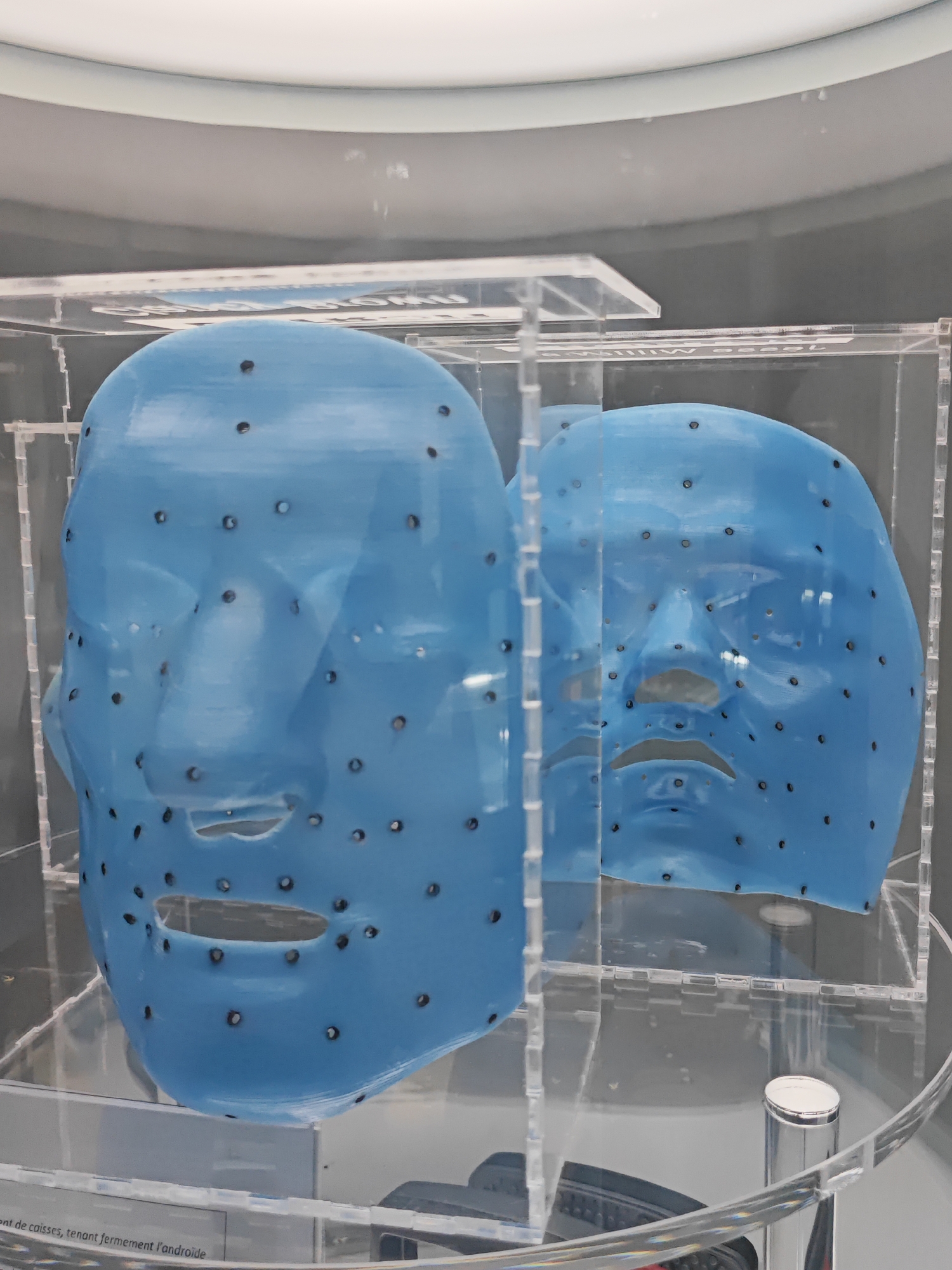 Two bright blue, mask-like objects shaped like human faces are displayed in clear acrylic cases. The masks are dotted with small black holes and feature cutouts for eyes, a nose, and a mouth. The masks are positioned one behind the other, with the front mask slightly to the left and appearing larger. The lighting is bright and diffused, casting a slight sheen on the masks. Below the masks, a reflective metallic cylinder stands upright. In the lower left corner of the image, text is partially visible on a white label, with some characters indicating French language. The overall environment suggests an indoor exhibition or display. The masks are located in Turin, Italy at the Museo Egizio.