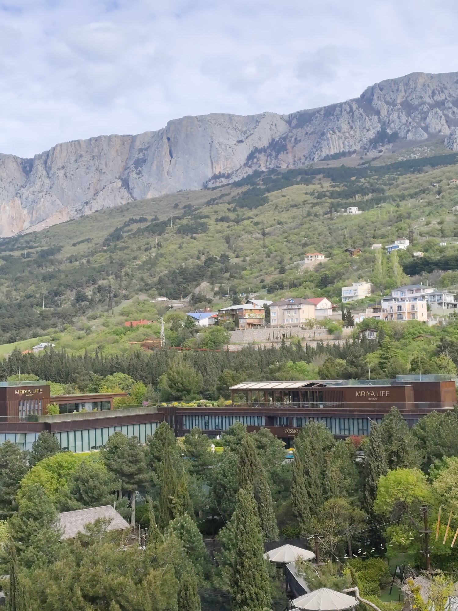 The image presents a daytime view of a mountainous region in the city of Симеизский поселковый совет, Ukraine. A mountain range with bare rock faces dominates the upper portion of the scene. The mountains are partially covered with green vegetation, interspersed with residential buildings on the slopes. In the midground, a modern building complex with "MRIYA LIFE INSTITUTE" emblazoned on the façade is visible, surrounded by lush trees. A smaller structure is partially obscured by trees in the foreground, along with elevated platforms and ropes suggesting an obstacle course or recreational area. The sky is overcast with clouds.