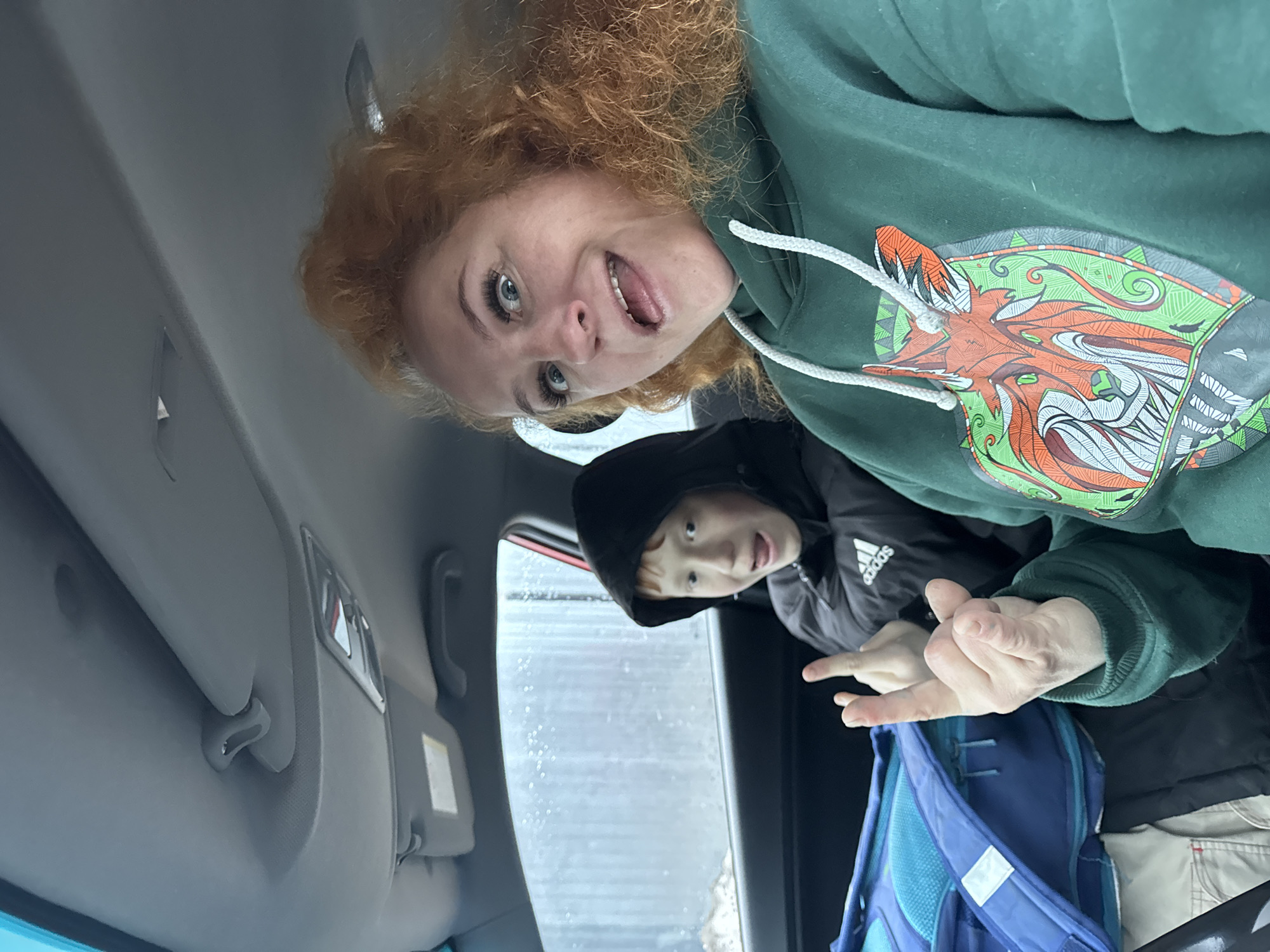 A woman and a boy are inside a vehicle, possibly a car, in Chaikovsky, Russia. The woman, with red curly hair, is wearing a green hoodie with a design of a wolf. She is looking at the camera with her mouth open. The boy, wearing a black Adidas hooded jacket, is also looking at the camera and gesturing with his fingers. Behind them, the window shows a blurred background, possibly indicating rainy or snowy weather. A blue backpack is visible near the boy. The interior of the car includes a sun visor.