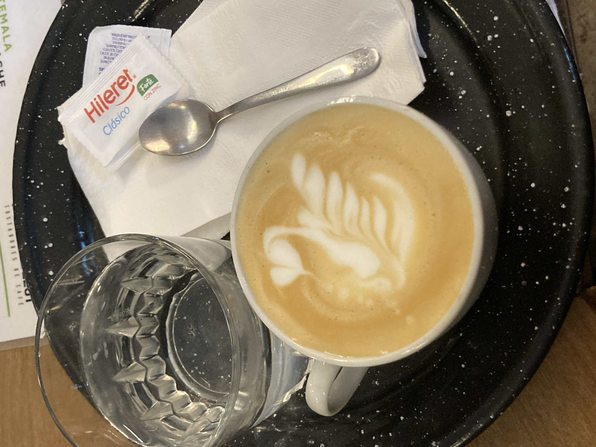 An overhead, close-up shot captures a coffee service presented on a speckled black tray. At the center, a white ceramic cup holds a light brown coffee beverage, likely a latte, adorned with intricate white latte art depicting a leaf or fern pattern. To the left of the coffee cup, a clear glass contains water, notable for its textured, possibly faceted, bottom.
A small silver spoon rests on a folded white napkin, positioned between the coffee cup and two small white packets. The packets are identified by the brand "Hileret." with the word "Clásico" in blue and "Forte CON ZINC" in green. Smaller text on one packet also reads "INDUSTRIA ARGENTINA," indicating its origin. Additional partial text from the packets includes "AZANTE," "JANTIENE," "FO DE SONG," "ONO, SULFATO," "AQLIITINANTE," "NP: 19-067355," and "19-000161."
A fragment of what appears to be a menu or paper is visible on the far left, with the words "GUATEMALA" and "BRES DE CAFE" partially legible. The speckled black tray provides a contrasting background for the items. The scene suggests a relaxed setting, likely a cafe or restaurant, where coffee and water are being served.