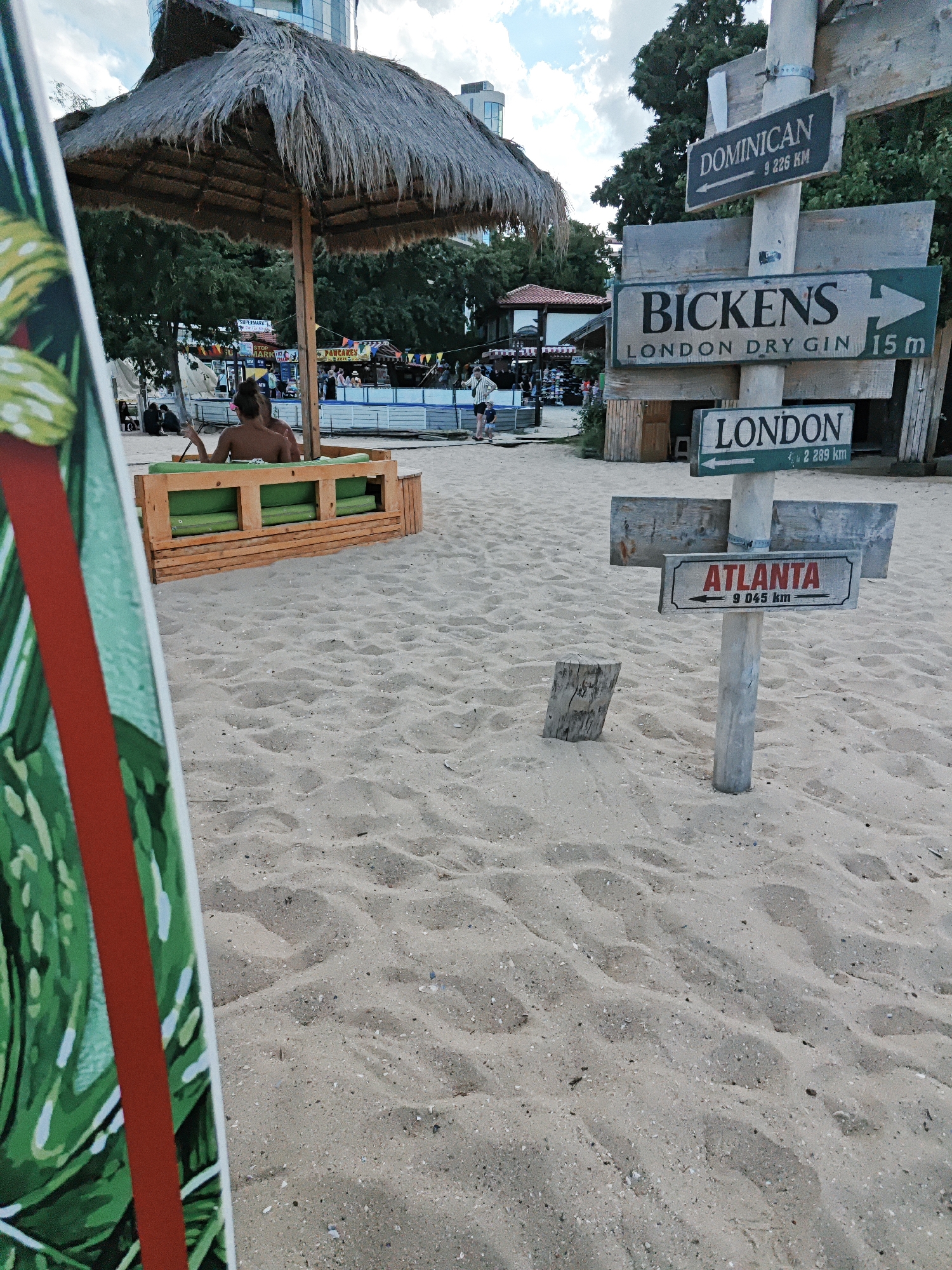 The image shows a sandy beach with a thatched umbrella in the background. Several directional signs are visible, indicating "DOMINICAN", "BICKENS LONDON DRY GIN", "LONDON", and "ATLANTA" with corresponding distances. In the mid-ground, a person is sitting on a wooden bench with green cushions. A partially visible surfboard with a green and red design is in the foreground on the left. The environment appears to be a beach resort or a coastal area, with trees and some structures in the distance. The sky is cloudy.