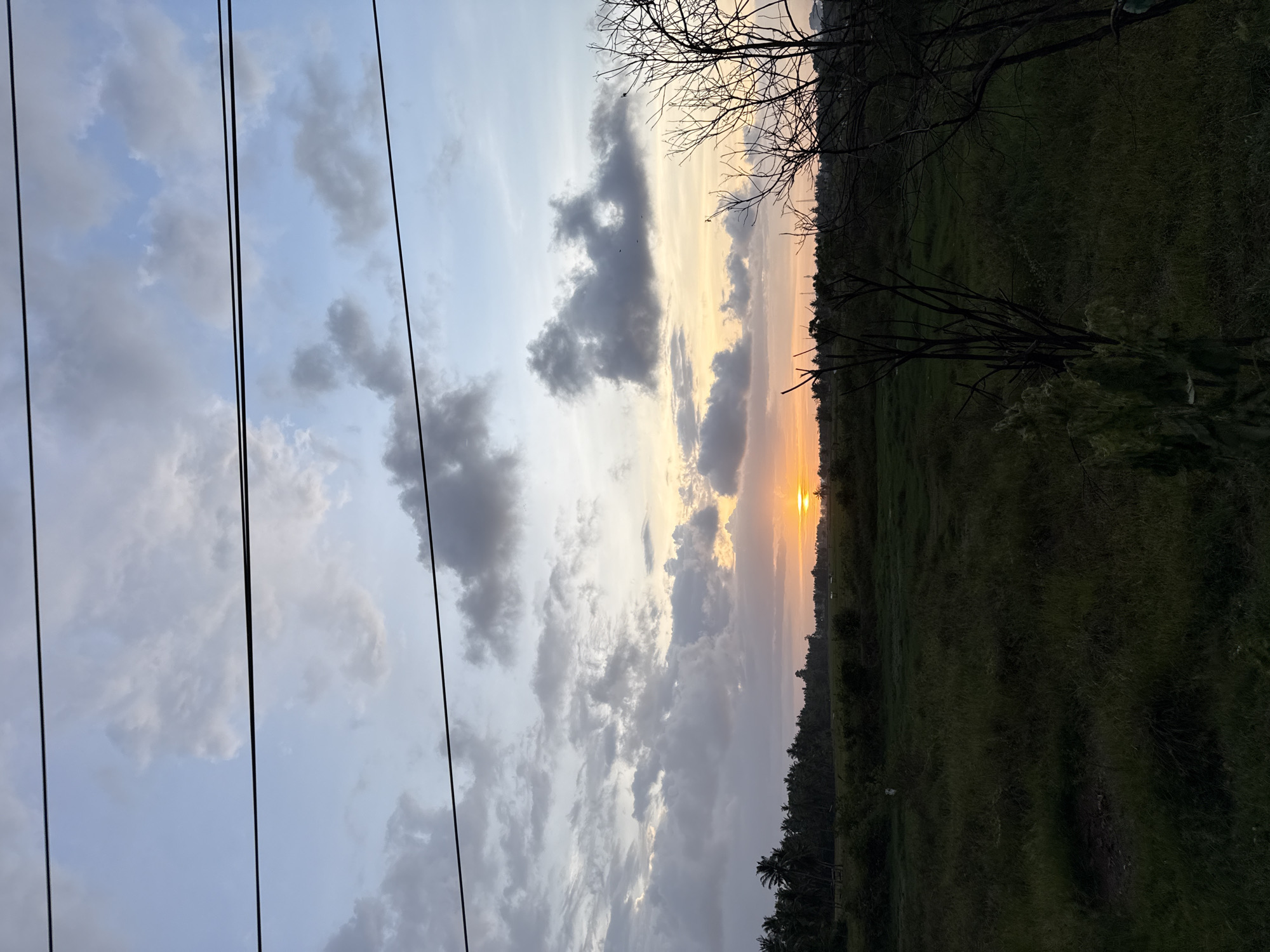 A vertical shot of the sky over Pulorungkom, Indonesia, displays a colorful sunset. The sky has blue sections interspersed with gray and white clouds. There are black power lines running vertically along the left side of the picture. The horizon is a thin line of orange and yellow where the sun is setting. The silhouettes of trees and vegetation are visible at the bottom of the image. Some bare tree branches are at the top right.