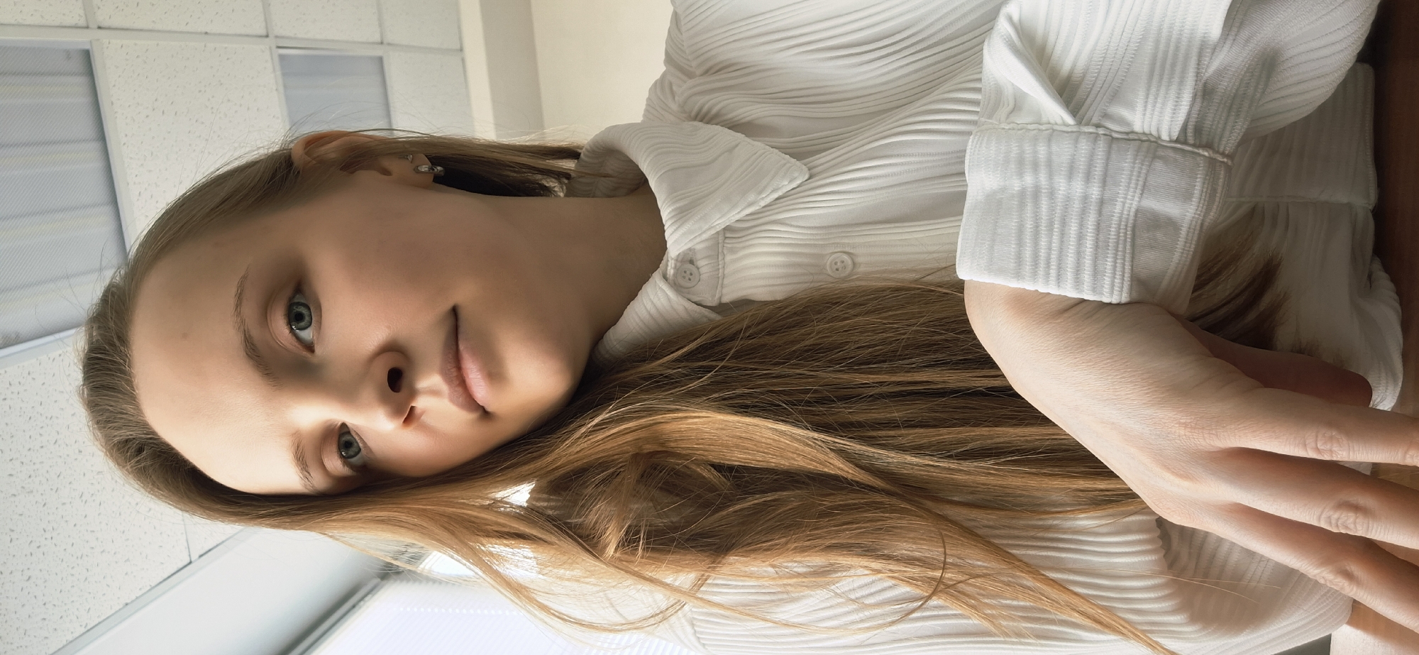 A woman with light skin, light brown hair, and blue eyes is the primary subject of the image. She is wearing a white, textured button-up shirt. Her hair is spread across the shirt. Her hand is also visible. The background shows a ceiling with square panels and a window. The image location and city cannot be determined from the visual information provided.