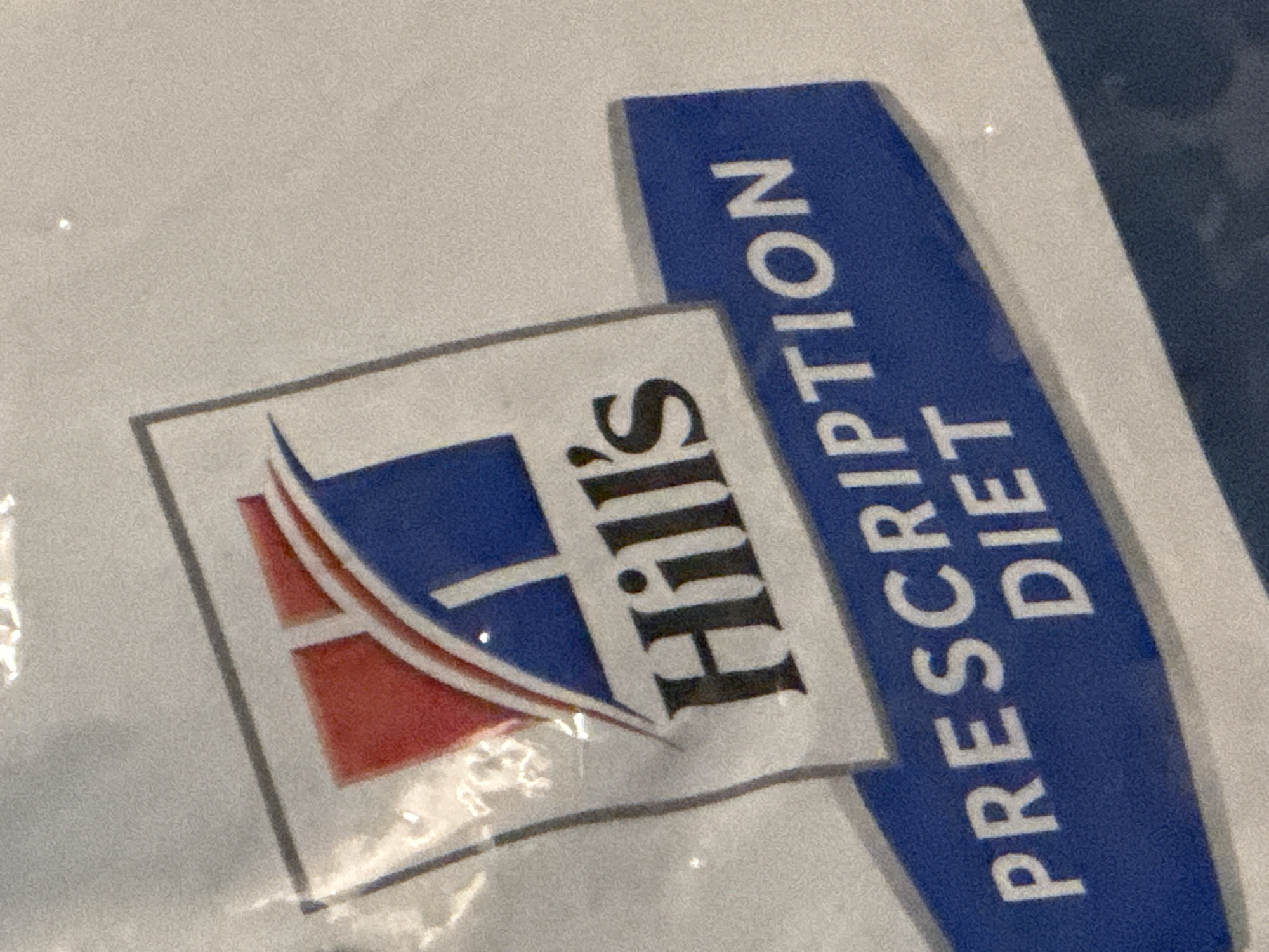 A close-up, angled shot shows a portion of a bag with the Hill's Prescription Diet logo. The logo features a stylized "H" in blue and white within a grey border, with red accents. Below the logo, the words "PRESCRIPTION" and "DIET" are printed in white capital letters on a dark blue background. The overall impression is of product packaging.
