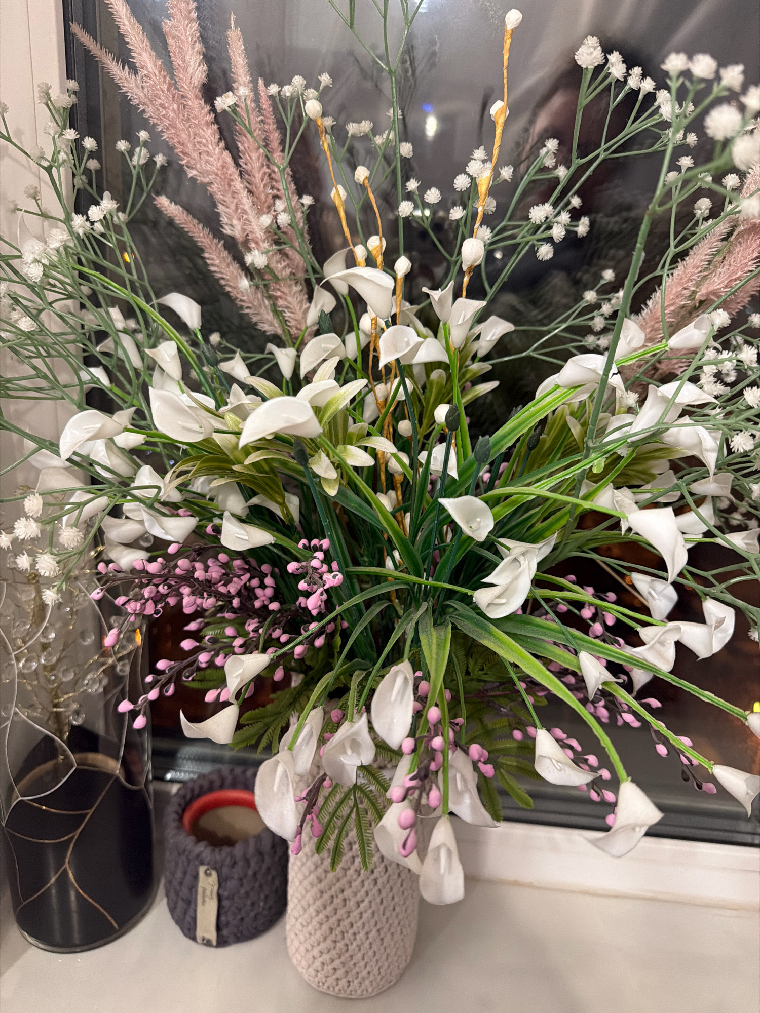A close-up, eye-level shot shows an artificial floral arrangement featuring white calla lilies, pink spray and light pink feathery plumes, and small white flowers resembling baby's breath. The flowers are arranged in a beige crocheted vase. To the left of the vase, a dark grey crocheted pot with a red interior holds a plant. Next to that, a black vase with gold line details contains a string of lights. The arrangement sits on a white surface, possibly a windowsill, with a blurred background suggesting a window and a dark interior. The scene is located on a windowsill in Vniissok, Russia.