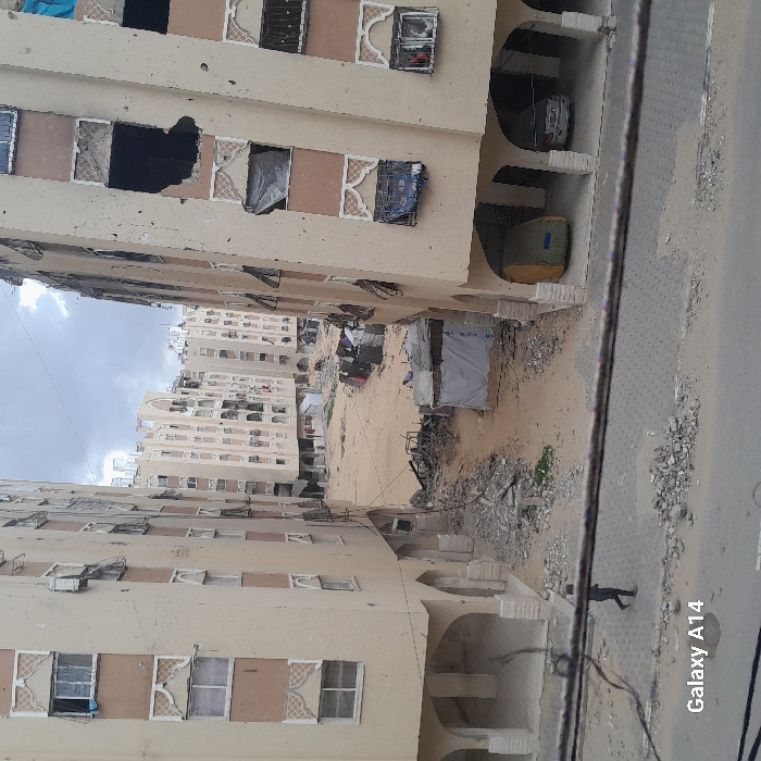 The image depicts a devastated urban landscape in Khan Yunis, Palestinian Territory, characterized by extensive structural damage to residential buildings.
In the foreground, a paved street strewn with concrete rubble and debris runs horizontally. A single individual, dressed in dark clothing, walks from left to right on this street. Above the street, dark overhead wires stretch across the scene.
Two multi-story, light-beige residential buildings frame an open area beyond the street. The building on the left shows numerous broken windows, some covered with white plastic or fabric. Its lower level features arched ground-floor passages. The building on the right exhibits severe damage, including a large, dark, gaping hole on an upper floor and multiple broken windows, many covered with dark tarpaulins. Beneath this building, two arched carports are visible; one contains a light-colored sedan, and the other houses a large object covered in a yellowish-green tarp.
Between these two foreground structures, a wide, open dirt and sand expanse is filled with piles of rubble, twisted metal, and discarded materials. Several small, makeshift shelters constructed from tarpaulin and various debris are scattered across this area. Further back, additional multi-story residential buildings, also appearing damaged with numerous broken windows, line the distant background. The sky is overcast with visible clouds. The overall scene conveys widespread destruction and hardship.