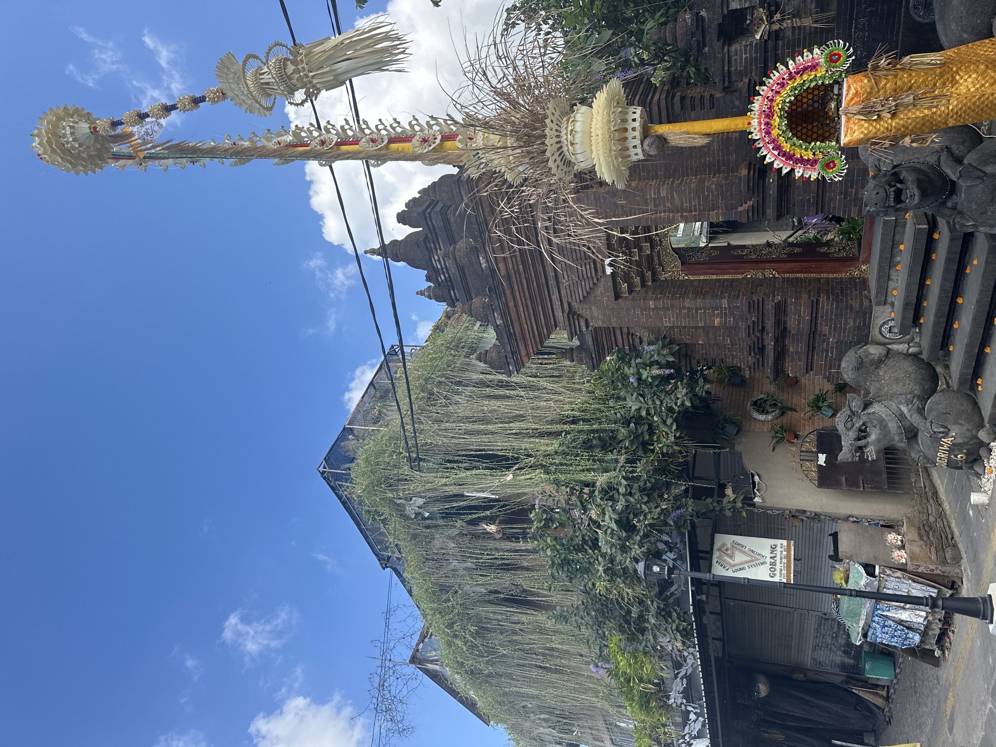 The image depicts a street scene in the Ubud District of Indonesia, likely during the day given the bright sunlight and clear blue sky with scattered clouds. The dominant subjects are elaborate decorations, possibly for a Balinese ceremony or festival, suspended high against the sky. These decorations appear to be made of woven palm leaves and colorful flowers, arranged in intricate patterns.
Beneath the decorations, a brick building with a tiered roof structure is visible, possibly a temple or traditional Balinese architecture. Lush green foliage, including trailing plants, cascades down the sides of the building, suggesting a humid, tropical climate.
In the foreground, a street-level view reveals more details. A lamppost stands prominently. There are piles of what appears to be discarded items or materials, including fabrics and bags, indicating a potentially busy or perhaps less organized area of the street. Next to these items, a sign is attached to a lamppost which reads "GOBANG SOUND SYSTEMS" and "FABIA LIGHTING." Further to the right, two stone sculptures, one resembling a mythical creature and another a human-like figure, are visible. The sculpture of the human-like figure has text inscribed on its base that appears to be "SUGIRIWA." Small offerings of flowers are scattered around the base of the sculptures. The overall impression is a vibrant cultural setting with a mix of traditional elements and everyday street life.