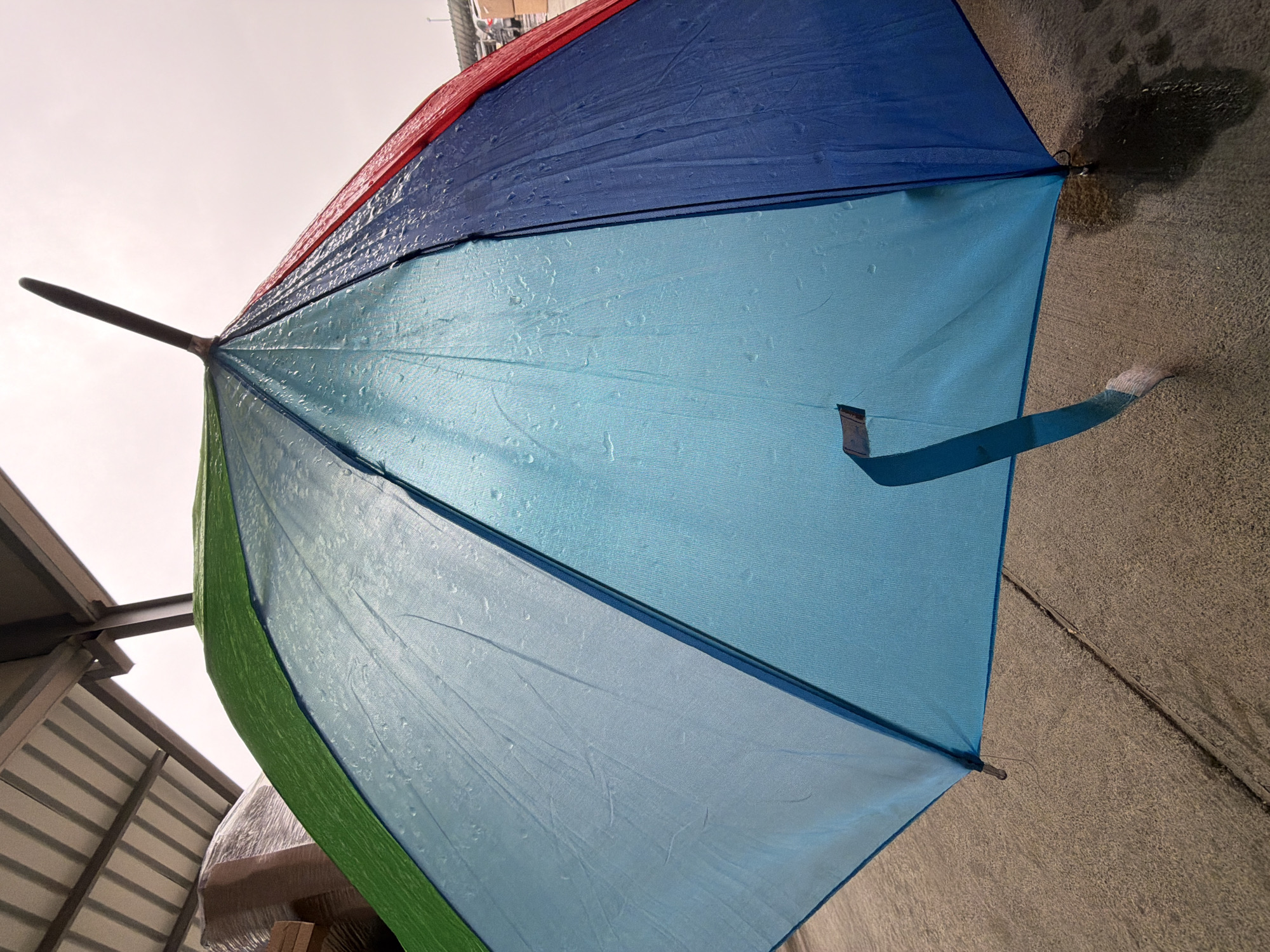 The image shows a wet, partially open, multi-colored umbrella with red, blue, light blue, and green panels. Water droplets are visible on the umbrella's surface. The umbrella's shaft extends to the left. A blue strap hangs from one of the panels. The background consists of a grey concrete surface. Part of a building structure with metal beams and corrugated panels is visible on the left side. The sky is overcast. The location is not explicitly identifiable as Parvenets, Bulgaria from the image alone.