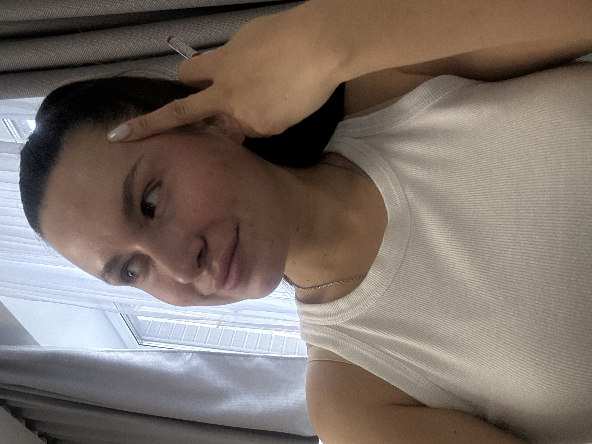 A person with fair skin, dark hair pulled back, wearing a white ribbed tank top, is indoors. The person is looking at the camera. Their right hand is raised near their forehead, with a thin red and silver object held between the fingers. A delicate silver necklace is visible around the person's neck. A window with white blinds and sheer white curtains is visible in the background. A gray fabric is behind the person and below the window.
The location is not clear from the image and there is no indication it is in the city of Odintsovo, Russia.