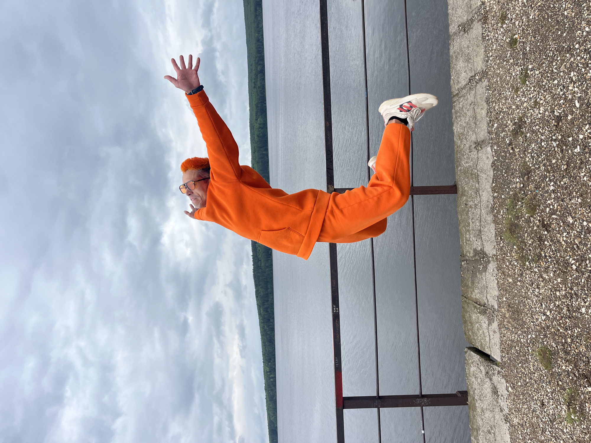 A man with orange hair, wearing an orange tracksuit and white sneakers, is posed horizontally in the air with arms raised against a backdrop of water, sky, and a concrete structure. His feet appear to be supported by a metal rail. The location is the city of Chaikovsky, Russia. Behind the man is a body of water with a wooded shoreline on the opposite side. The sky is overcast. The concrete structure is a pier or platform.