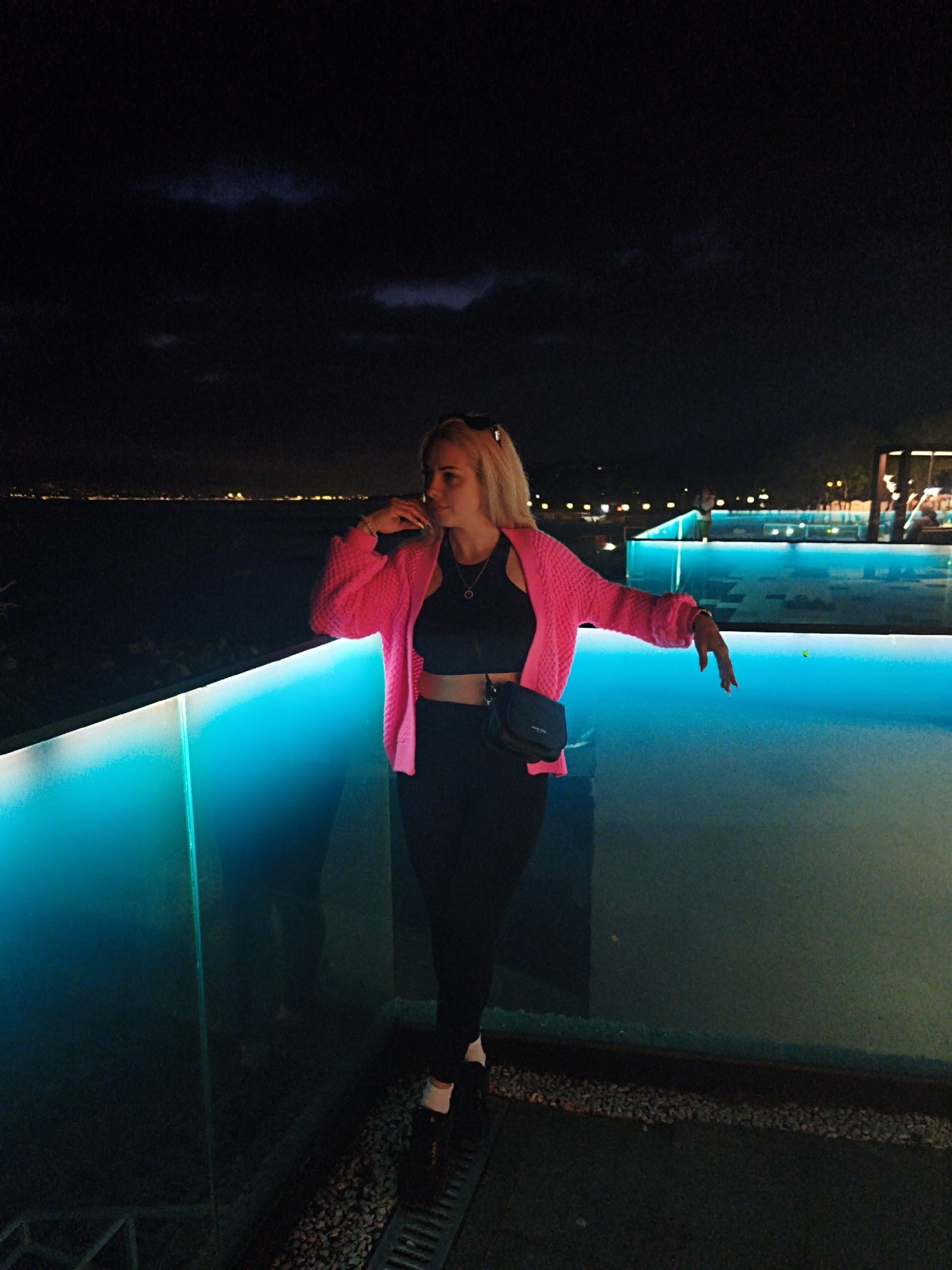 A woman stands on a balcony overlooking an illuminated pool at night in Novorossiysk, Russia. She wears a pink cardigan over a black crop top and black pants, with black shoes and white socks. She is holding a glass, possibly drinking, and has a black crossbody bag. The balcony has a glass railing with blue LED lighting. In the background, there is a body of water with city lights and a dark sky with clouds. The ground of the balcony consists of small rocks next to a metal drain.