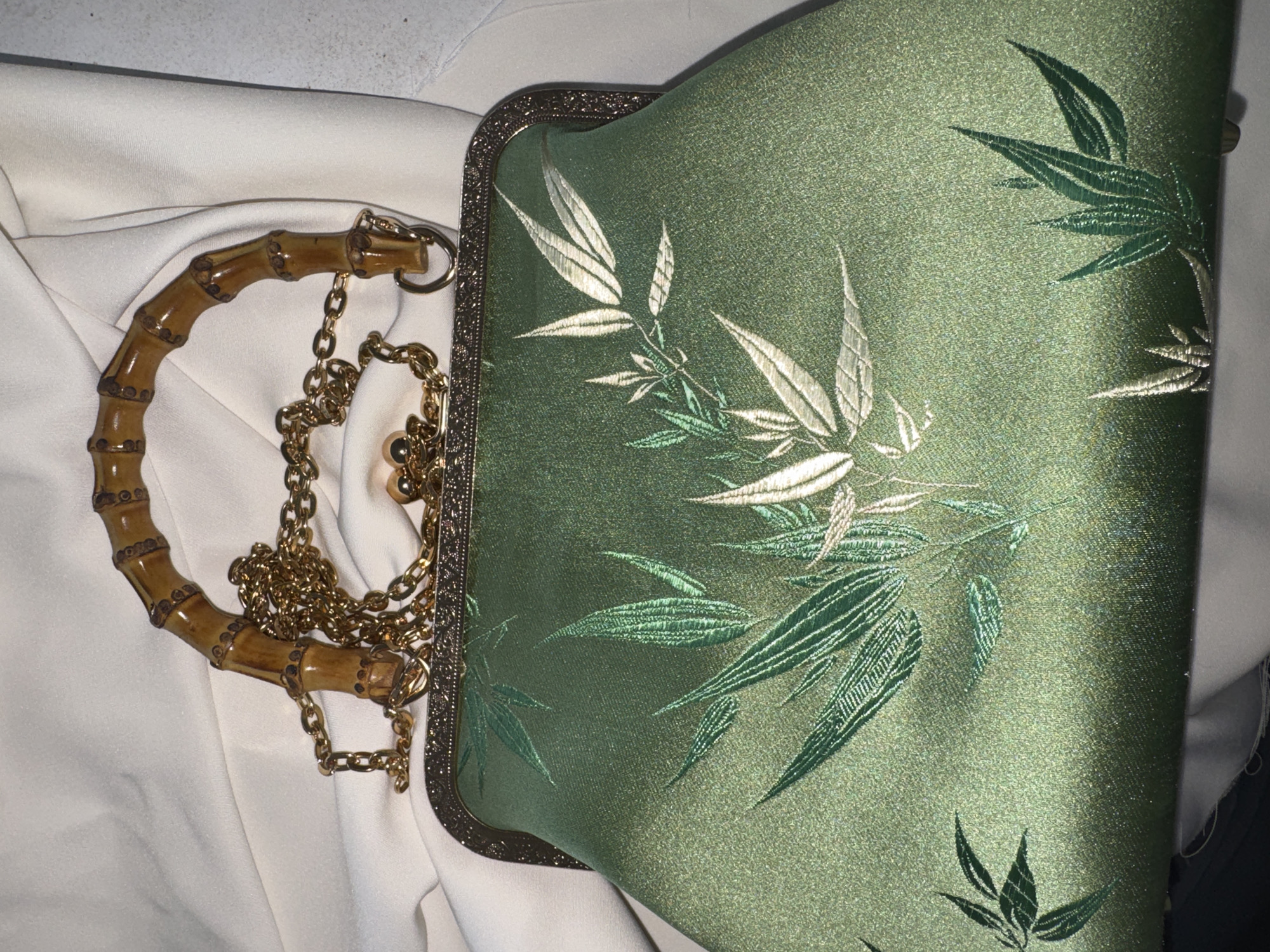 A green silk clutch purse with a bamboo handle and a gold chain strap is displayed on a white, draped fabric. The purse is decorated with an embroidered pattern of bamboo leaves in various shades of green and metallic silver. The frame of the purse is ornate and gold-colored, with a clasp closure. The background is out of focus, suggesting a shallow depth of field. The lighting is soft, with some shadows present.
