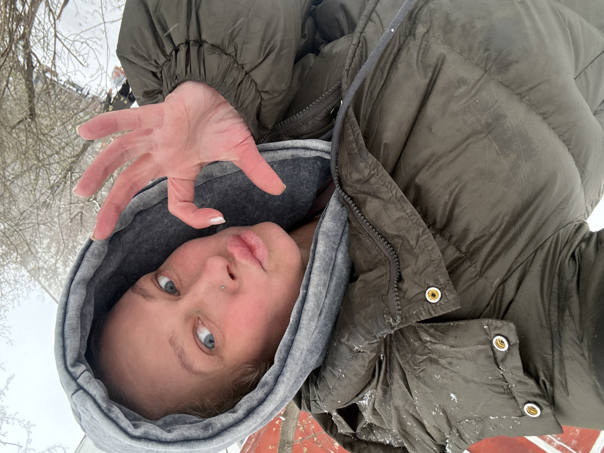 This media file is a rotated portrait-oriented selfie featuring a young woman in an outdoor, snowy environment, likely in Perm, Russia, as per the provided context. The image is rotated 90 degrees clockwise, showing her head towards the left and her body extending towards the right.
The central subject is a woman looking directly at the camera with light blue or grey eyes. Her lips are slightly parted and puckered. She has a small silver stud piercing on her left nostril. Her hair, though mostly covered, appears light in color. She is bundled in winter attire, wearing a grey hooded sweatshirt with the hood pulled up, under a dark olive or khaki green puffer jacket. The jacket shows visible signs of snow or ice flakes, suggesting cold weather. Her right hand is raised with fingers spread towards the camera, with light-colored nail polish visible on her fingertips.
The background is blurred but clearly depicts a winter scene. Bare tree branches, heavily coated in frost or snow, are visible against an overcast sky. The overall impression is a cold, possibly freezing, daytime environment. A hint of red or orange is visible in the bottom right corner of the image (when oriented upright), which could be part of a structure or ground covering. The activity is a self-portrait taken in a snowy setting. No visible text appears in the image.