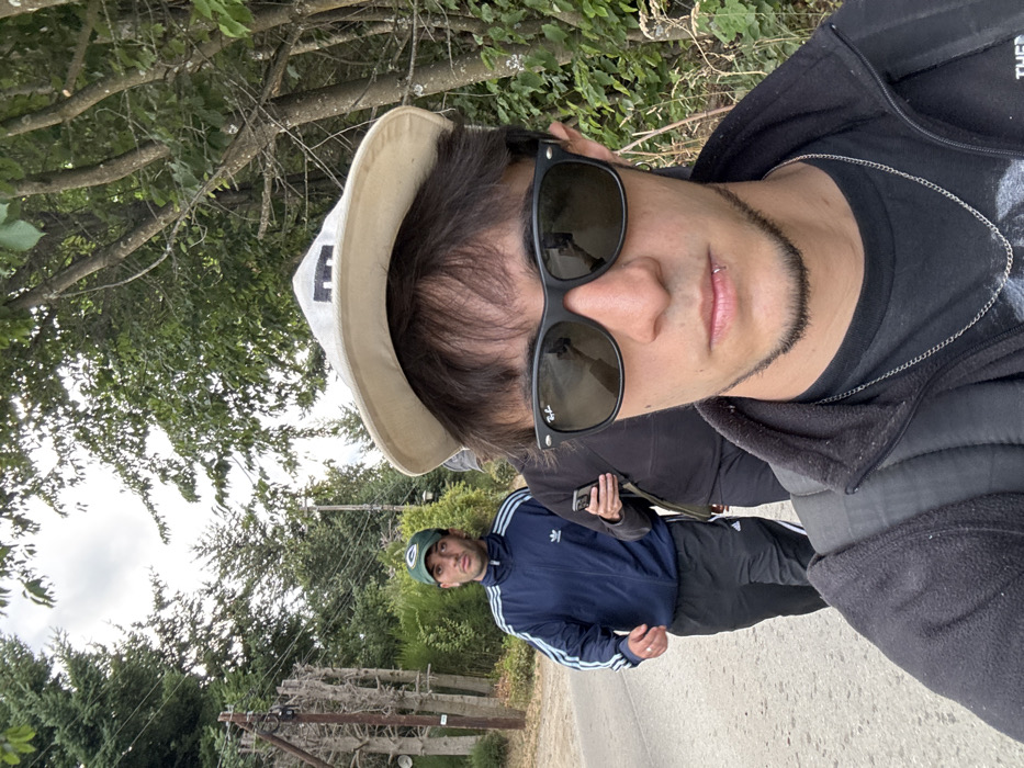 Two men on a path are shown. The person closest to the camera wears sunglasses, a hat with the letter "E" on the side, and a black hooded jacket over a black t-shirt. A silver chain hangs around his neck. Behind him is another man in a green baseball cap and a blue and white Adidas jacket and black pants. They are walking on a path bordered by greenery and trees. A fence can be seen in the distance.
