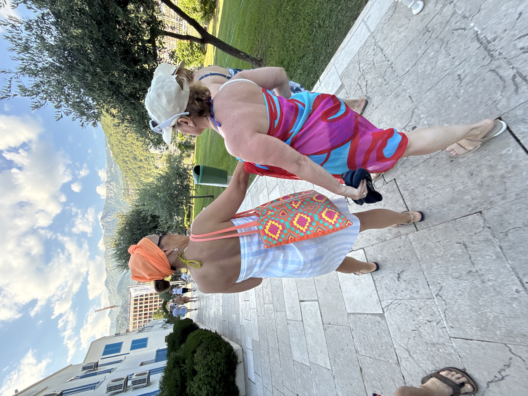 The image shows two women walking along a paved walkway. The woman in the foreground is wearing an orange headscarf, sunglasses, a white and blue striped dress, and a colorful patterned bag. The woman behind her is wearing a white baseball cap, sunglasses, and a vibrant pink, blue, and red sarong. The background features lush green trees, a grassy area, and a mountainous landscape under a partly cloudy sky. In the distance, there are buildings and a construction crane. The scene appears to be set in a warm, sunny environment, likely during the daytime. The overall mood seems relaxed and casual, suggesting a holiday or vacation setting. The orientation of the image is rotated 90 degrees counterclockwise.