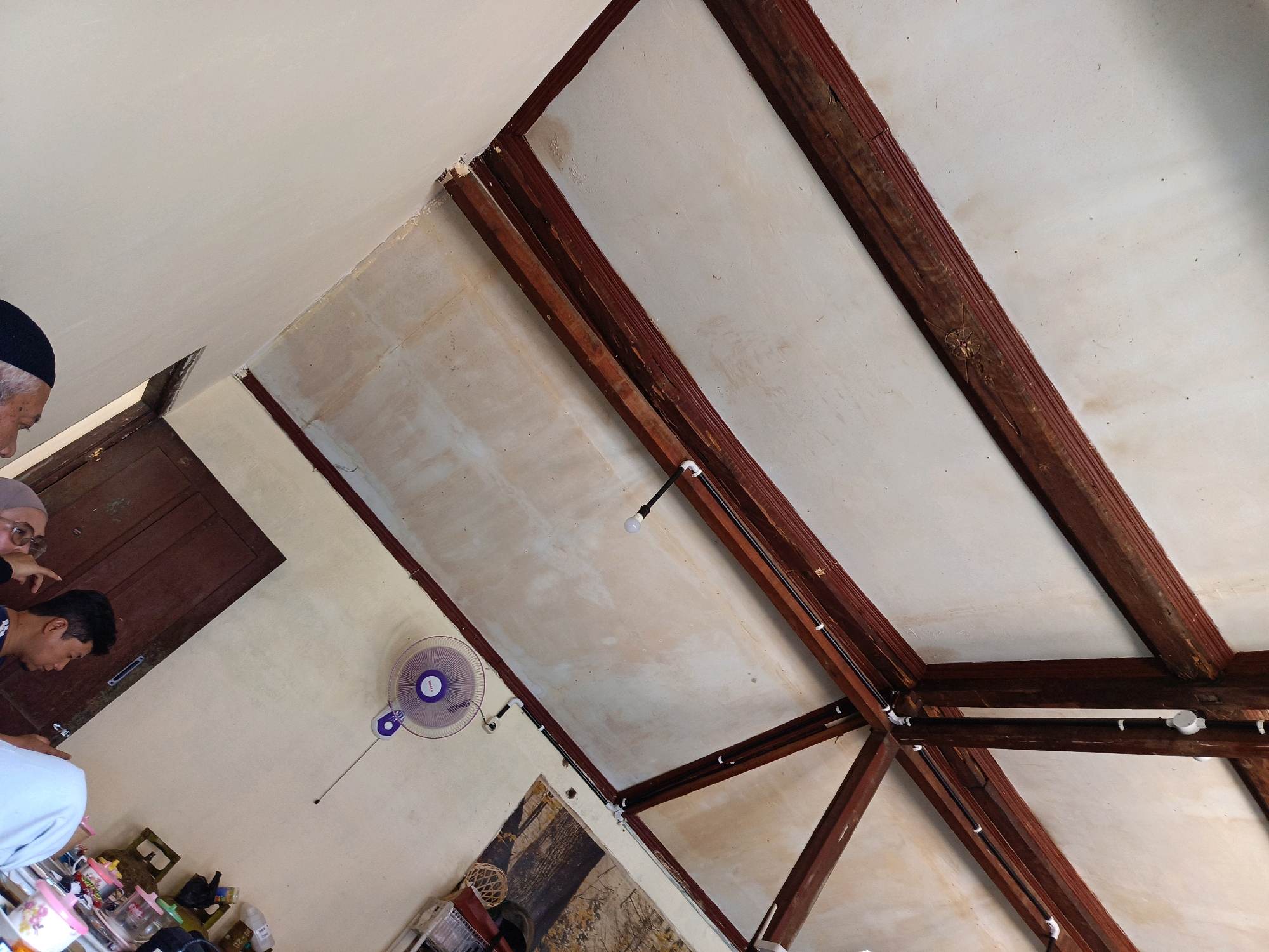 The image depicts an interior scene, likely in Kebonagung, Indonesia, with a focus on a rustic wooden ceiling structure. The ceiling is supported by thick, dark brown wooden beams, some of which are distressed and show signs of age. The ceiling panels themselves are a light, off-white color, with some discoloration and texture visible.
In the lower left portion of the image, a section of a light-colored wall is visible, with a dark wooden door positioned on the left edge. A purple oscillating wall fan is mounted on this wall. Below the fan and to the right, there are various objects on a surface, including containers and what appears to be a basket.
Three individuals are partially visible in the upper left of the frame. The person on the far left is wearing a black skullcap and is looking downwards. Next to them, another person wearing glasses and a headscarf is pointing to the right. A third person, a young man with dark hair, is also looking downwards. They are positioned near the wooden door.
The lighting suggests an indoor environment, possibly during the day, with natural light and some artificial lighting from the visible ceiling fixtures. The overall impression is of a simple, perhaps older, building with exposed structural elements.