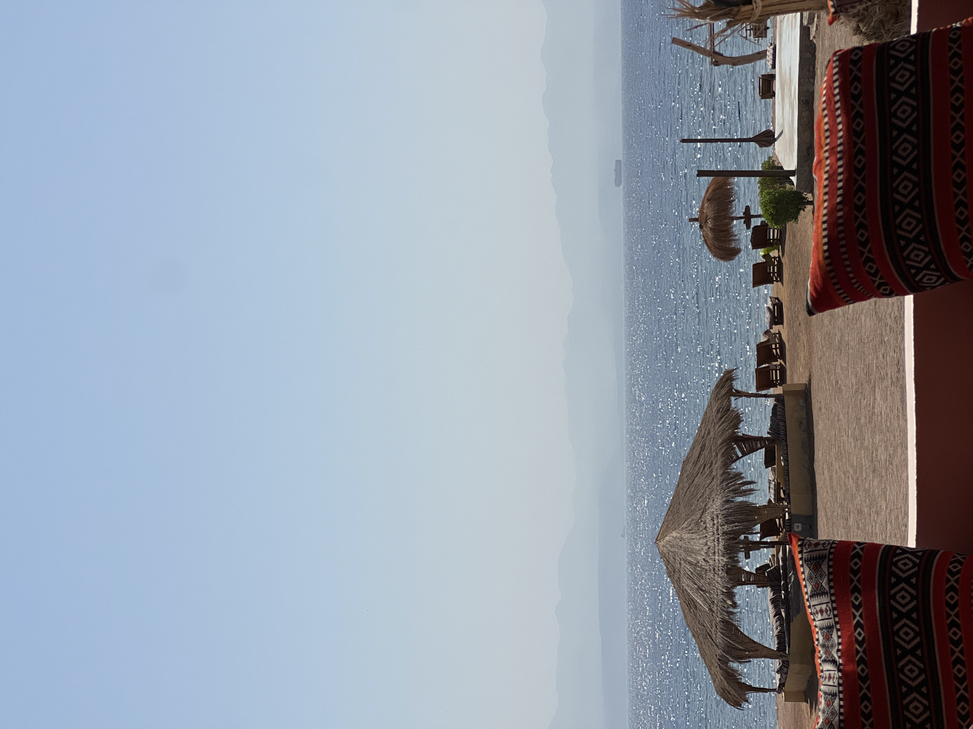 A bright, sunny, vertical, medium-shot image shows a beach scene in Nueiba, Egypt. In the foreground, a red and black patterned cushion is angled towards the viewer. The cushion is on a reddish-brown surface, possibly a low wall. In the mid-ground, a straw-thatched gazebo with seating inside is located on a sandy beach. Several lounge chairs are visible behind the gazebo, and to the right, there's a smaller thatched umbrella. The sea, with its surface sparkling in the sunlight, stretches across the middle of the image, and in the far distance, hazy blue mountains are visible on the horizon. A cargo ship is depicted sailing on the sea. The sky is a pale, cloudless blue.