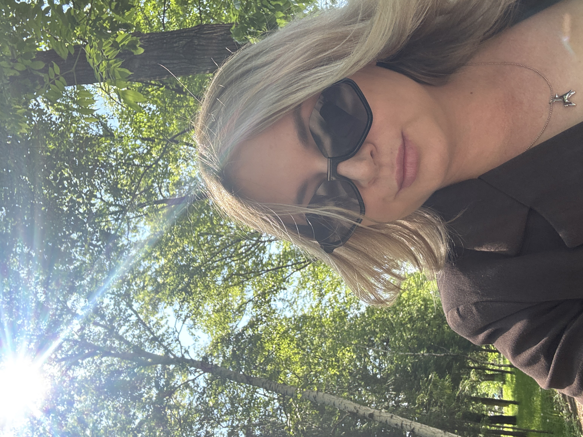 A woman with blonde hair and sunglasses is taking a selfie outdoors. She is wearing a dark brown blazer and a silver necklace with a small dog pendant. The background shows a bright, sunny day with green trees and some birch trees. The image is shot from a low angle, looking up at the woman. The sunlight creates lens flare in the upper left corner of the image. The setting appears to be a park or wooded area. It is not possible to determine the specific location within the city of Birobidzhan, Russia, from the provided image.