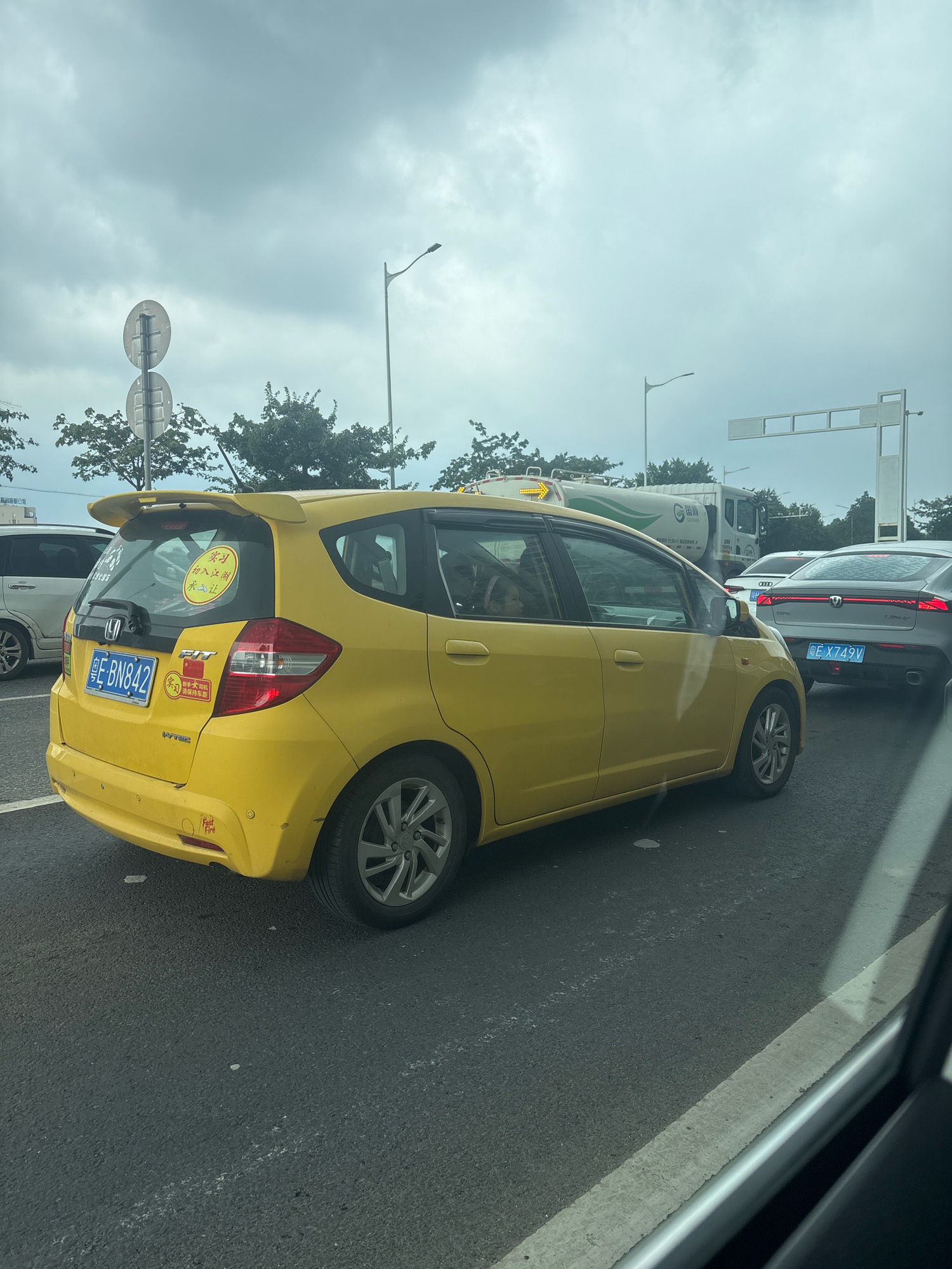 The image shows a yellow Honda Fit taxi, license plate E-BN842, driving on a road in Sanshui District, China. The taxi has a yellow sticker on its rear window with Chinese characters and a circular red logo. Other vehicles, including a white SUV and a silver sedan with license plate E-X749V, are visible on the road. A large truck with a white tank is also in the background. The sky is overcast. The scene appears to be a traffic jam.