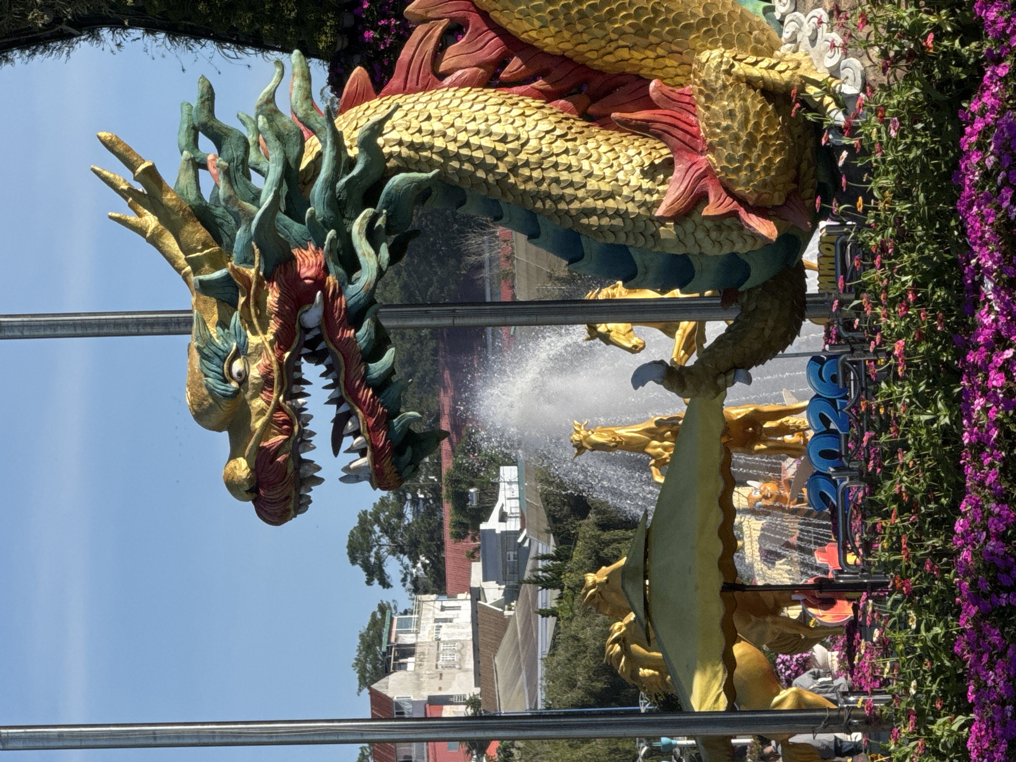 A large, golden dragon sculpture with green and red accents is the focal point of this outdoor scene, set against a bright blue sky. The dragon's head, with its mouth open in a roar, is prominently displayed on the right side of the frame. Behind the dragon, a fountain sprays water upwards, partially obscuring golden horse sculptures and an yellow umbrella. In the foreground, a bed of vibrant purple and pink flowers blooms profusely. A row of blue numbers, appearing to read "2026", is visible behind the flowers. In the background, buildings with red and white rooftops and lush green trees are visible, suggesting an urban or semi-urban setting. The image appears to be taken in Phường Xuân Hương - Đà Lạt, Vietnam, given the architectural style and foliage. There are no discernible people in the image, but a glimpse of a person in dark clothing is visible on the far left edge.