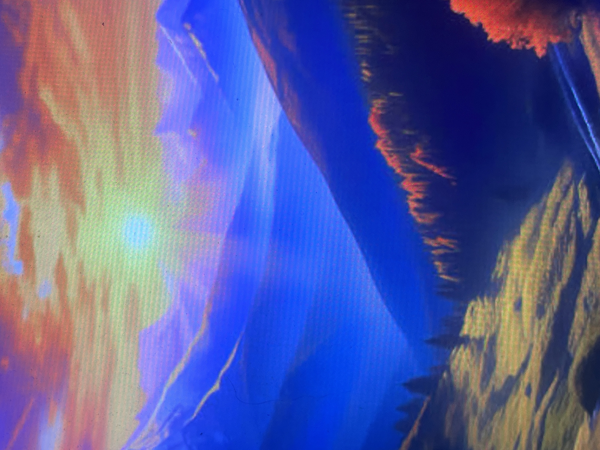 The image is a photograph of a digital display, exhibiting a visible grid pattern and screen artifacts. It depicts a stylized, panoramic mountainous landscape at either sunrise or sunset, characterized by a highly saturated and vibrant color palette.
The upper portion of the scene features a sky with warm-toned clouds, primarily orange and red, blending into purple and blue hues. A radiant, greenish-yellow spherical light source, consistent with a sun, is centrally positioned in the upper-middle, emitting a distinct glow and faint rays.
Multiple layers of mountains extend across the midground and background, displaying deep blue and purple colorations, indicating atmospheric perspective. These mountains form steep slopes and valleys.
In the mid-to-foreground, the terrain includes forests and rolling hills. On the right side, a dense forest covers a steep slope, with trees exhibiting strikingly bright red and orange foliage, suggestive of autumn. On the left side, rolling, grassy hills or pastures are visible, composed of muted green and yellow tones, with scattered dark green, conical trees. A winding, lighter path-like feature is present in the lower-left foreground.
The image contains no discernible human figures, animals, or specific man-made structures. The landscape is generic in its depiction of natural elements and lacks any unique geographical markers, landmarks, or contextual clues that would forensically link it to a specific real-world location, including the city of Zelenyi Hai, Ukraine. The scene appears to be an artistic rendering or illustration of an idealized natural environment.