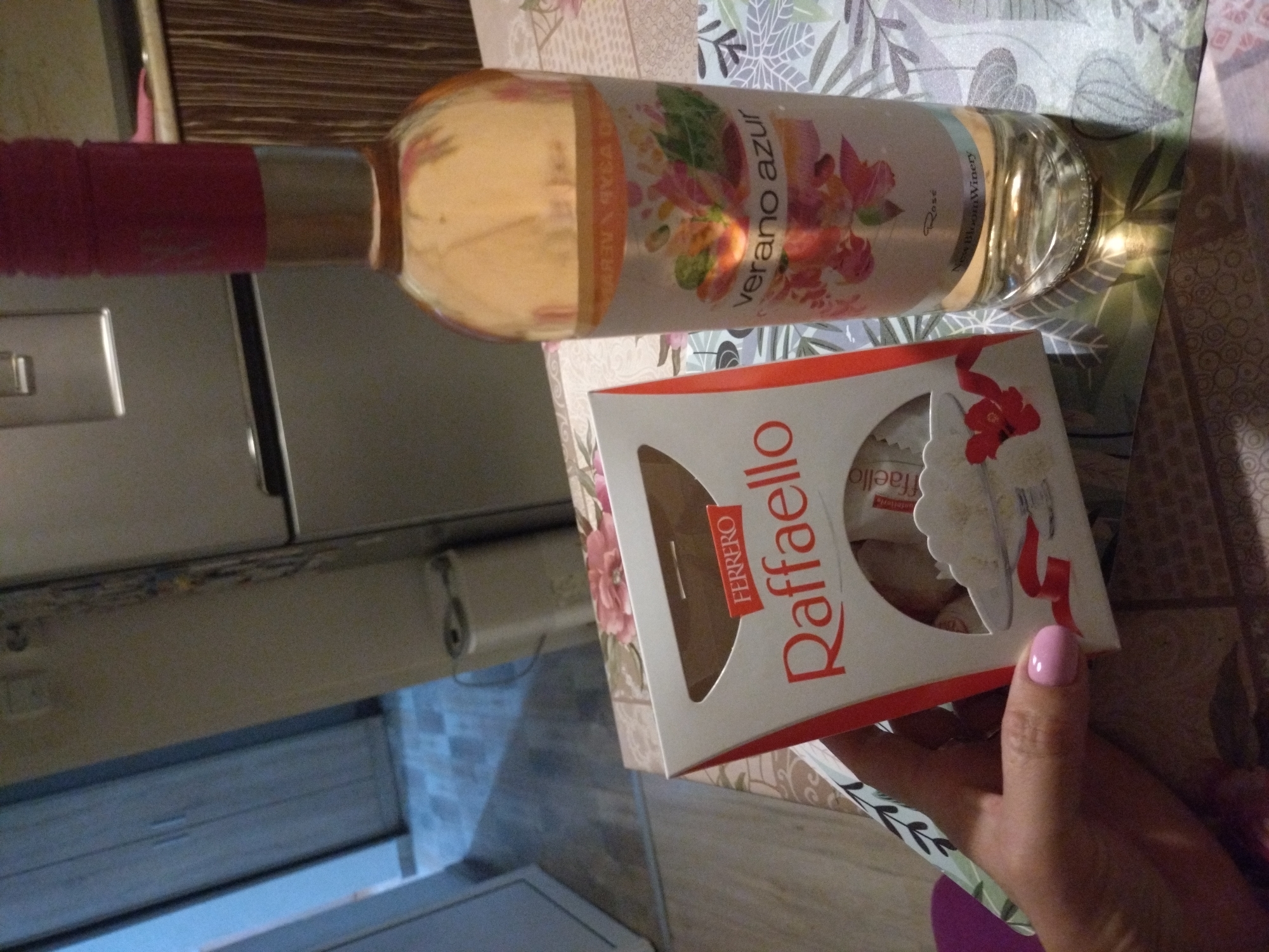 The image presents an indoor scene, consistent with a domestic environment in the city of Stara Zagora, Bulgaria, as specified by the user; however, no visual elements within the image itself confirm this specific geographical location. In the foreground, a light-skinned hand with pink-painted fingernails is positioned on the lower left, holding a white and red cardboard box of "FERRERO Raffaello" candies. The box's top is open, revealing multiple white-wrapped confections inside. Adjacent to the Raffaello box, a clear glass bottle containing a light pink liquid, labeled "verano azur" Rosé from "New Bloom Winery," stands upright. The bottle is sealed with a red foil cap. These items are situated on a patterned surface, appearing to be a tablecloth or placemat adorned with floral and leaf designs in muted green, pink, and brown tones. The background depicts a typical household interior, featuring a silver two-door refrigerator on the right, next to dark wood-grained cabinetry. To the left, a lighter-colored wooden door or wardrobe is visible, leading into an area with a light-colored, possibly tiled, floor. A small, light-toned household appliance is discernible on the floor in the mid-background. The lighting is artificial and diffuse, casting subtle shadows. No other individuals are present, and the sole action depicted is the hand holding the candy box.
