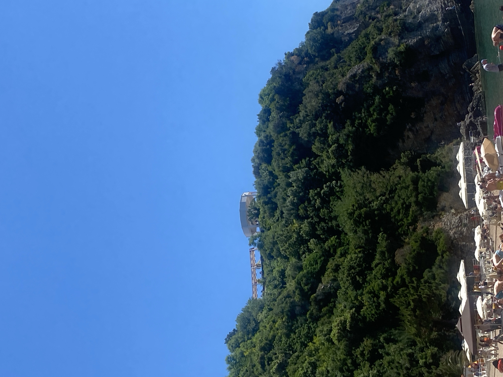 The image captures a scene in Budva, Montenegro, featuring a steep, green hillside dominating the view, with a clear blue sky in the upper left. A unique structure, possibly a viewing platform or part of an architectural installation, is situated atop the hill, accessible by a stairway. In the bottom right corner, a beach area is visible, populated with white umbrellas and sunbathers, suggesting a resort or recreational setting along the Montenegrin coast. The juxtaposition of the natural landscape with the man-made elements and the recreational activities indicates a popular tourist destination.