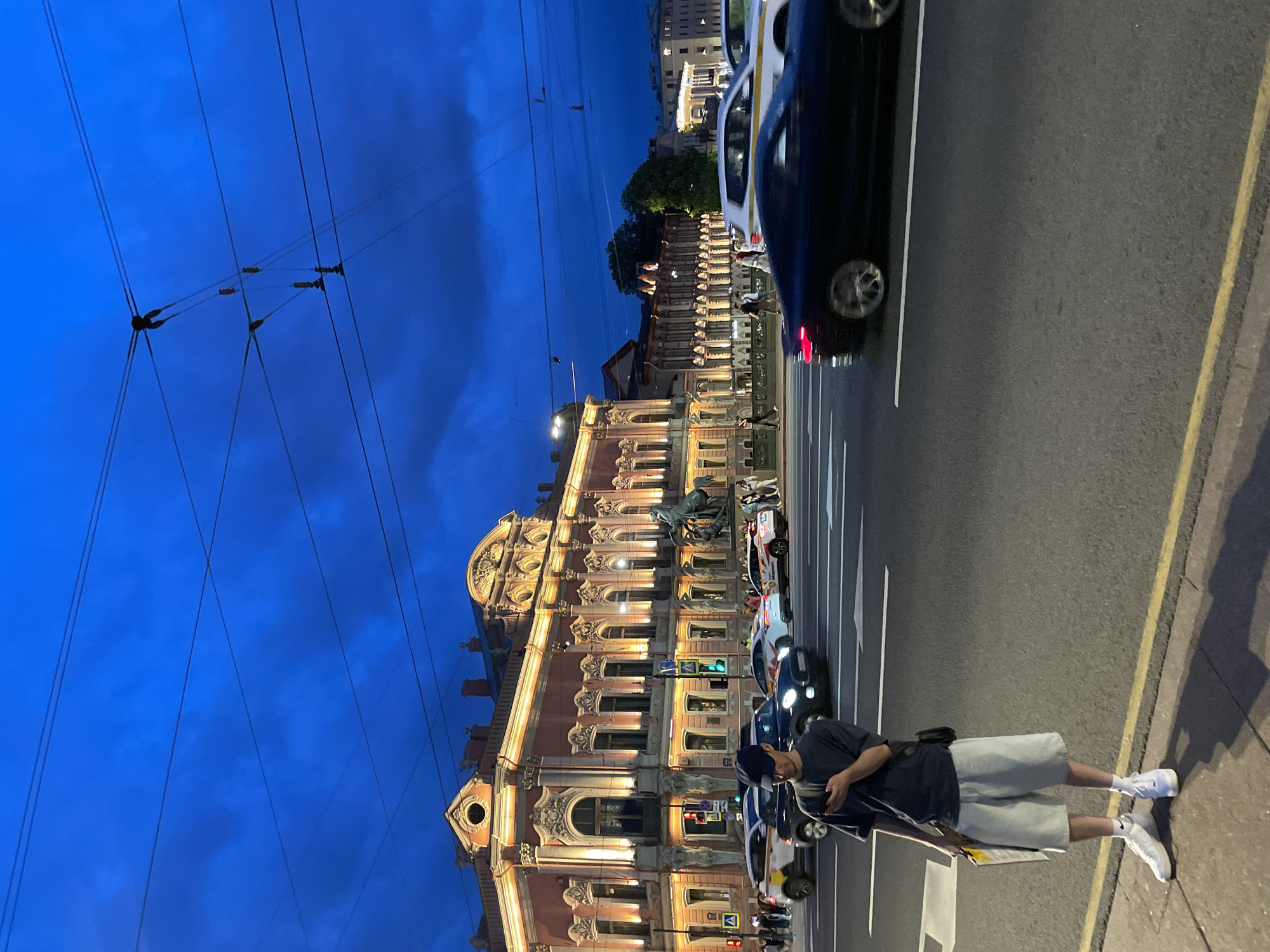 The image captures an evening scene on Nevsky Prospekt at the Anichkov Bridge in Saint Petersburg, Russia. The sky is a deep indigo blue with scattered clouds, crossed by overhead wires.
In the foreground, a young man stands on the edge of the sidewalk, facing slightly left. He wears a dark cap, dark t-shirt, light gray shorts, and white sneakers, with a black shoulder bag. He holds a smartphone in his right hand and a yellow item, possibly a folder, in his left. His shadow stretches across the asphalt.
Behind the man, a multi-lane street is active with traffic. Several cars are visible, including white taxis with yellow markings, and a dark car in the immediate foreground lane shows motion blur, indicating movement. Traffic lights are visible across the street.
To the left, dominating the midground, is the illuminated Beloselsky-Belozersky Palace. This grand, ornate building features a peach-colored facade with intricate architectural details, balconies, and numerous windows glowing with warm yellow light. Adjacent to the palace, one of the famous "Horse Tamers" equestrian statues from the Anichkov Bridge is partially visible. Further down the street to the right, other buildings and trees are partially visible, also illuminated by city lights, beneath the blue sky. Pedestrians can be seen walking on the sidewalks in the background.