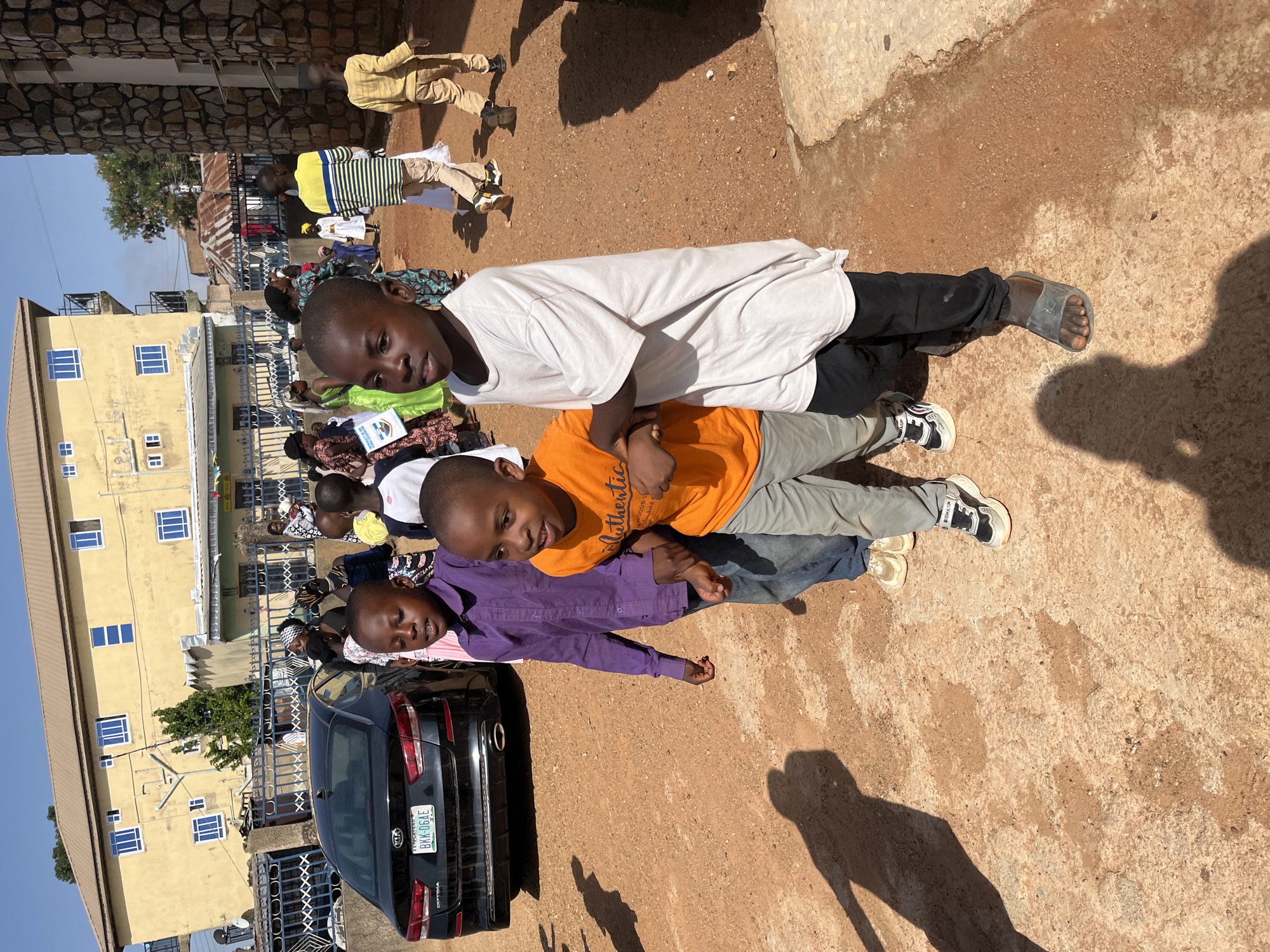 Three young boys lie in a stack on a dirt and concrete surface. The boy on top wears a white t-shirt and black pants, with grey sandals on his feet. The middle boy has an orange t-shirt and light grey pants, while the bottom boy wears a purple long-sleeved shirt and jeans. The boys are stacked head-to-toe in a line. To the left of the boys is a black KIA car with a license plate reading "BKK-06AE" from LE Plateau, Nigeria. Behind the car is a yellow building with blue-framed windows. Other people stand near the building, and more people are further away in the background. The sun casts a shadow on the ground. The location appears to be in or near Gura Top, Nigeria.