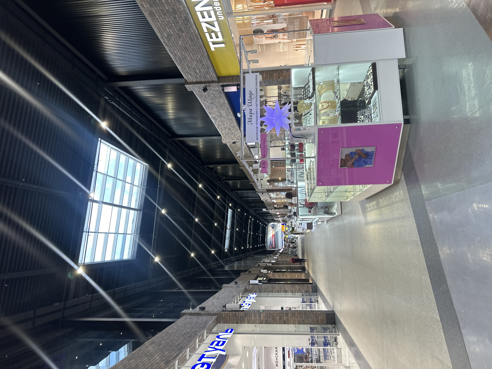 The image shows a long, indoor shopping mall corridor. The ceiling is high with a dark, industrial design featuring exposed beams and ductwork, illuminated by natural light filtering through a large skylight and multiple artificial lights. The walls of the corridor are lined with various retail stores, some with glass facades displaying merchandise. On the left, a store with a blue and white sign that reads "STYLE" (partially visible) and another with "ZHENSKAY" (women's) are visible. On the right, a store with a yellow sign that reads "TEZENIS" and "under..." is prominent. In the middle of the corridor, a small kiosk or display stand is set up, featuring jewelry and accessories. This stand has a pink and white structure with glass display cases showcasing necklaces, earrings, and bracelets. A light blue star-shaped decoration hangs above the kiosk. Several people are sparsely scattered throughout the mall, walking or browsing. The floor is a polished grey material. The overall impression is of a modern, well-lit shopping environment.