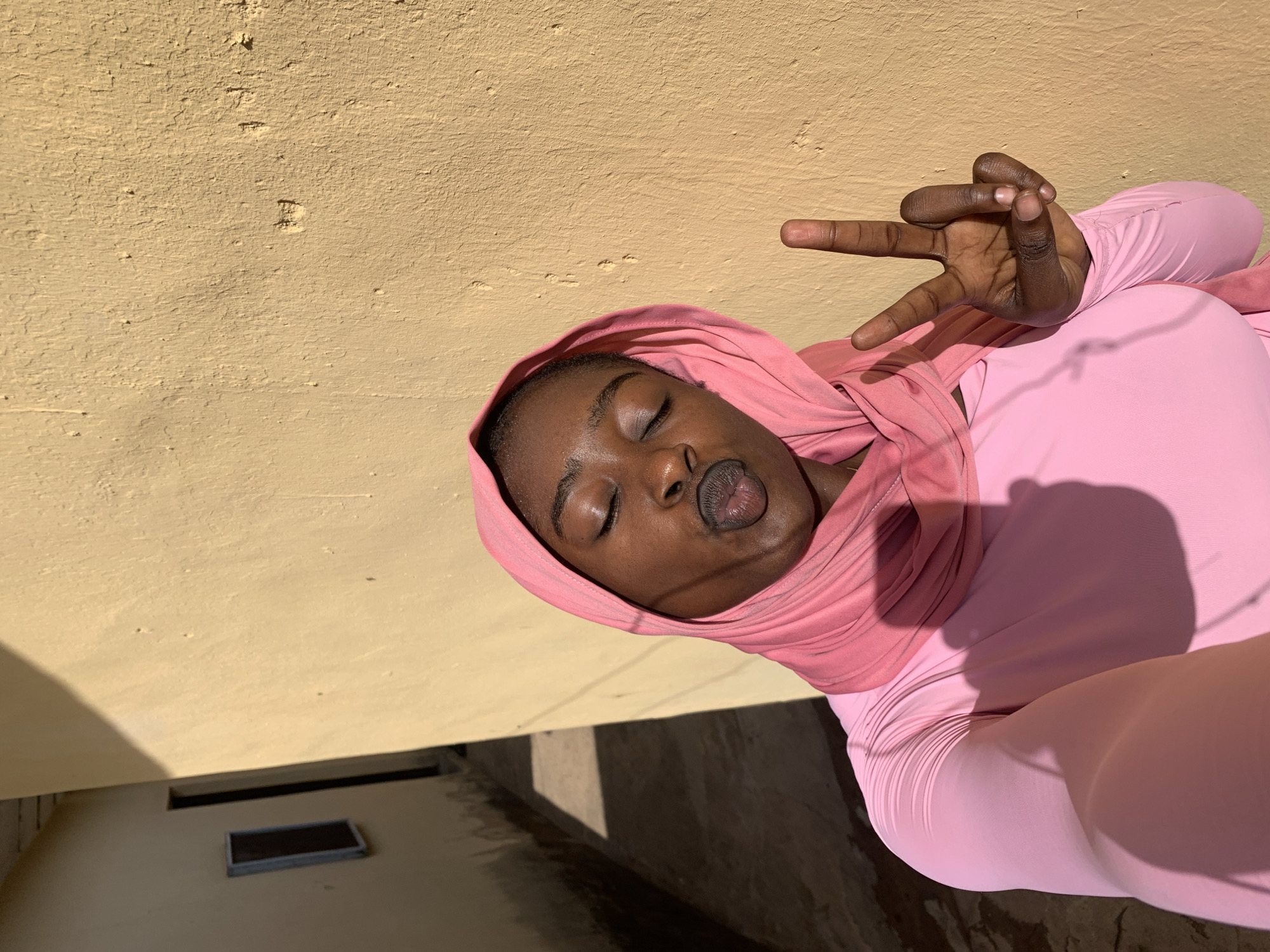 The image shows a person wearing a pink hooded garment, possibly a hijab, standing next to a light yellow stucco wall with visible wear. The person is making a hand gesture with two fingers extended. The location is indicated as Rafin Par, Nigeria. The background shows a portion of the ground with shadows and a dark rectangular opening.