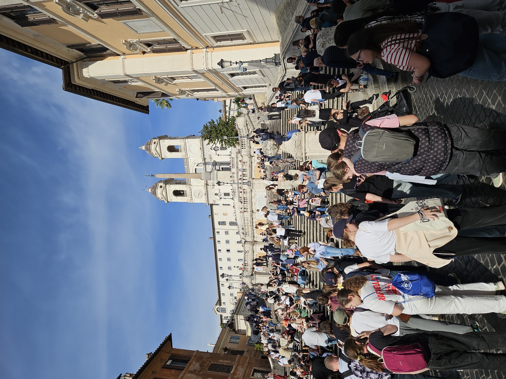 The image depicts a daytime scene at the Spanish Steps in Rome, Italy. The iconic staircase is filled with a large crowd of people, many of whom are sitting or lounging on the steps. Buildings flank the staircase, with architectural details visible. The sky is clear and blue. The Basilica di Trinità dei Monti is visible at the top of the Spanish Steps. People are wearing varied clothing, suggesting a mix of locals and tourists.