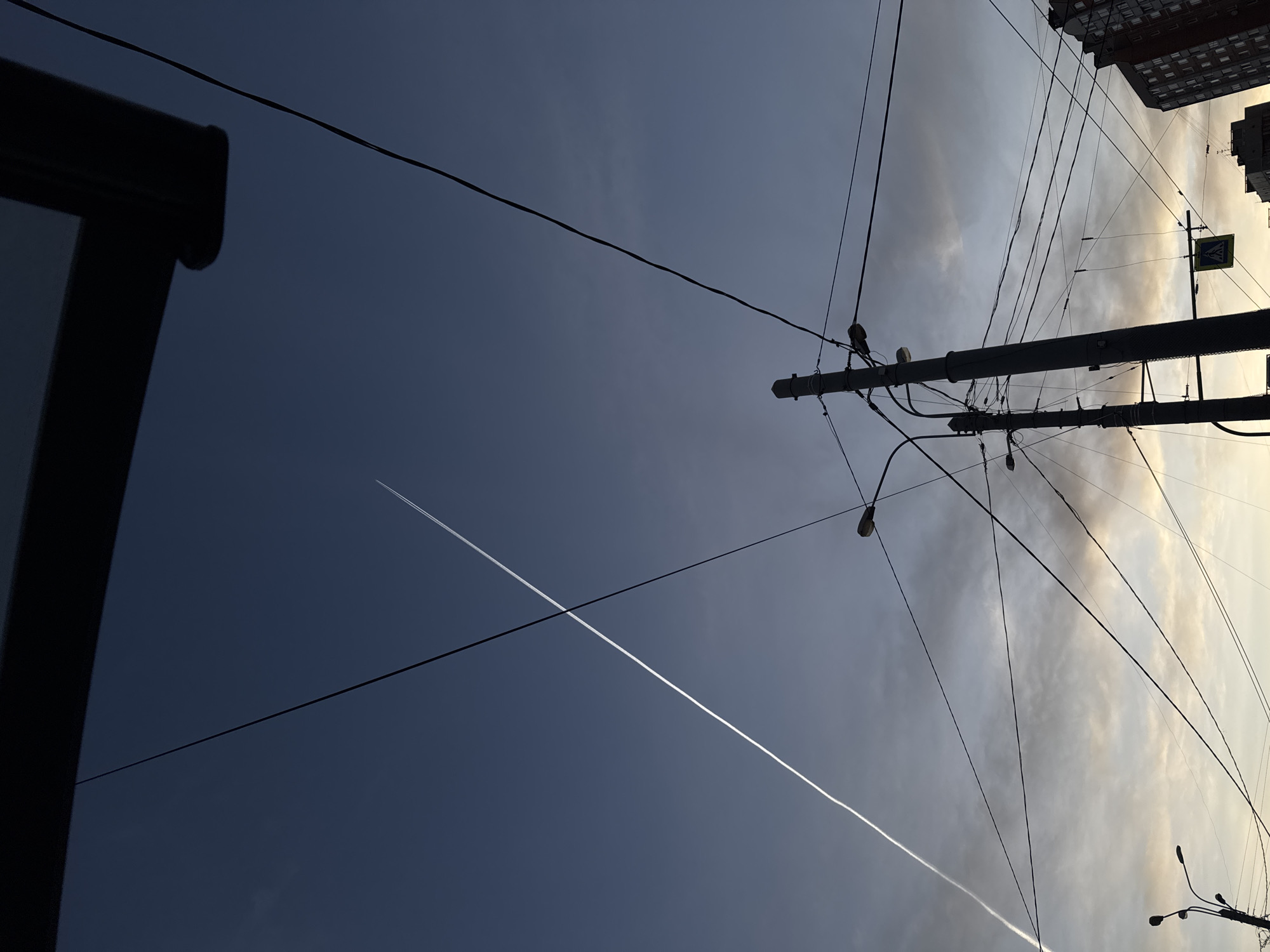 This dynamic image, captured from a low angle looking upwards, presents a striking urban sky punctuated by utility infrastructure and a luminous airplane contrail. The sky displays a distinct gradient, transitioning from a deep blue-grey on the left to a lighter, cloud-dappled, golden-grey on the right. A prominent, bright white contrail slices diagonally across the darker portion of the sky, intersecting with one of the numerous dark wires.
A central grey utility pole, heavily laden with a dense network of black electrical and communication wires, dominates the midground. These wires extend in various directions, forming intricate linear patterns against the sky. Multiple streetlights are attached to the pole and suspended from the wires. On the far right, the dark, silhouetted forms of multi-story buildings are visible against the brighter sky, suggesting they are backlit by a setting or rising sun. A dark, angular structure is partially visible on the far left edge of the frame, possibly part of another building or street fixture.
Attached to one of the utility poles on the right is a square yellow sign with a dark black symbol, identifiable as a standard warning sign for a pedestrian crossing. The strong contrasts, the sky's color gradient, and the silhouetted elements collectively suggest either early morning or late afternoon light, characteristic of dusk or dawn. The weather appears partly cloudy. The scene depicts a bustling urban environment, consistent with the provided context of Saint Petersburg, Russia. No people are visible in the frame.