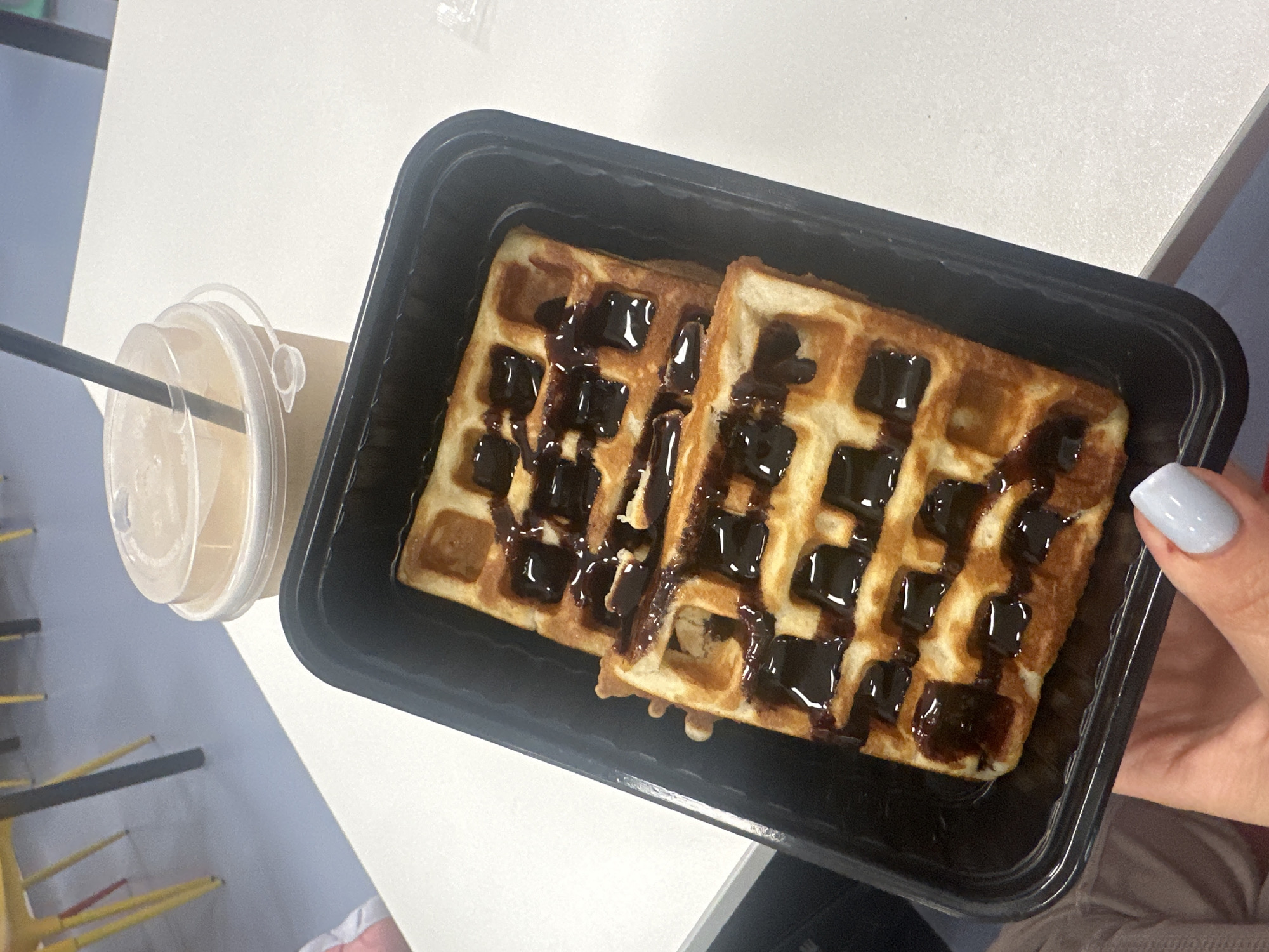 A top-down view of a black plastic food container filled with two waffles topped with chocolate sauce sits on a white surface next to a partially visible coffee cup with a straw. A hand with a light blue fingernail holds the container. The environment includes a chair and a hint of clothing worn by the person holding the container. There is no explicit evidence to confirm the location is Khimki, Russia.