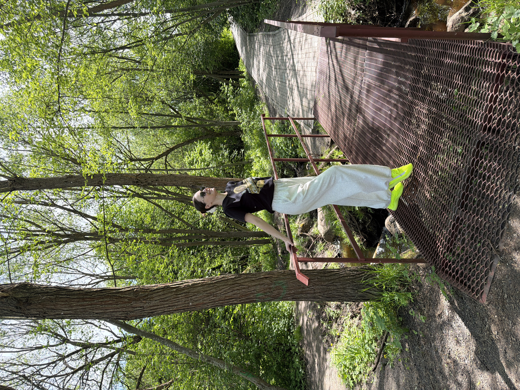 A photograph depicts a person standing on a metal bridge in a wooded area. The person is wearing a black t-shirt, light-colored pants, and bright neon green shoes. They are also wearing sunglasses and appear to be looking upwards. The bridge has a mesh-like surface and red metal railings. Trees surround the bridge, with some branches and leaves visible overhead. A path is visible on one side of the bridge, and there's a stream or water body underneath it. The lighting suggests it is daytime, with shadows cast on the ground and bridge. The location is an area with dense vegetation, implying a park or natural setting.