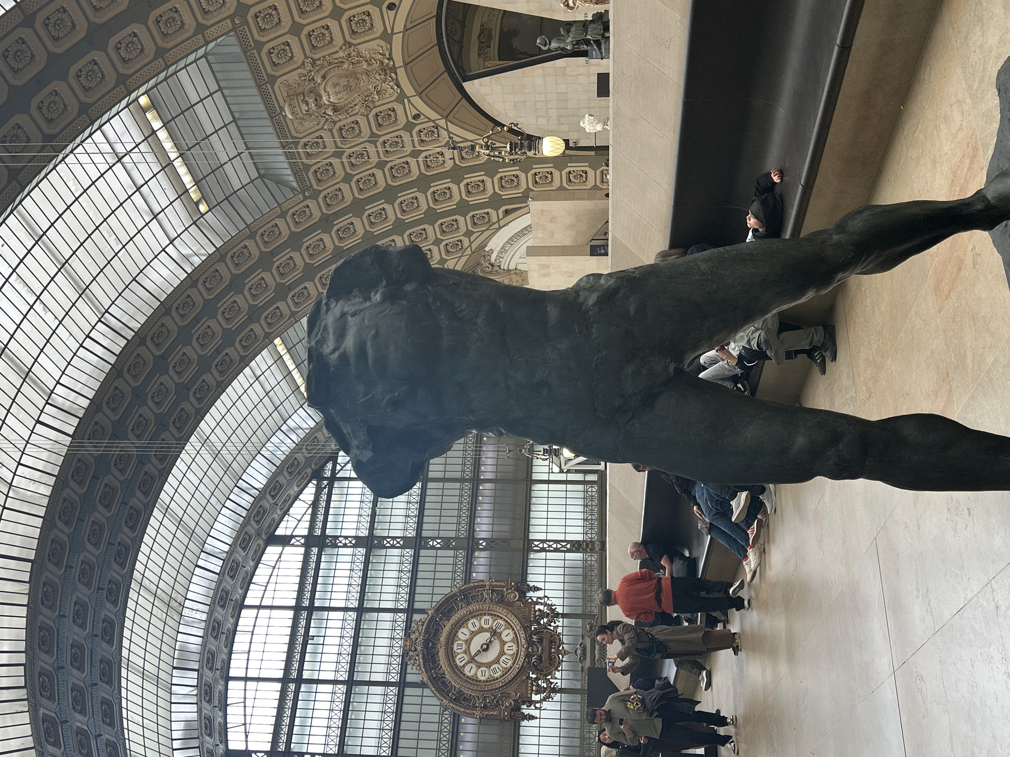 The image is taken inside the Musée d'Orsay in Paris, France. A large bronze statue of a nude male figure dominates the foreground. The statue is positioned to the left of the image's vertical center and is angled with its lower body towards the bottom right. Behind the statue and to the right, a group of people is observed, many of whom appear to be museum visitors. Some are standing, and others are seated. Further in the background, an elaborate clock with Roman numerals hangs in front of a large window with a grid pattern. Above, the museum's architecture is visible, featuring a curved, patterned ceiling with decorative elements. Light enters through the glass roof.