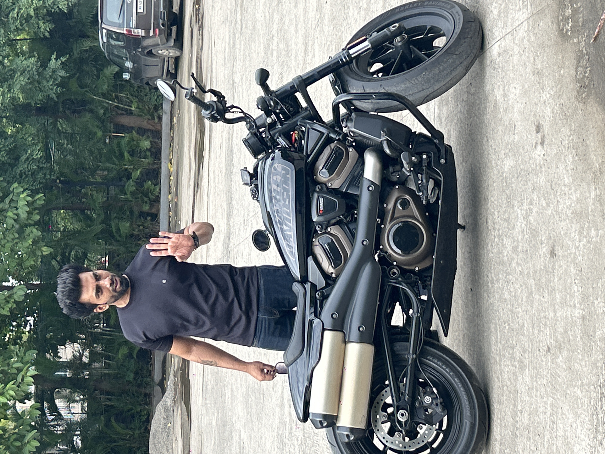 In Pune, India, a man with dark, spiky hair and a beard stands beside a black Harley-Davidson motorcycle. He is wearing a black t-shirt and blue jeans. His right hand is raised, palm facing the viewer, in a gesture that appears to be a stop signal. He is holding a pair of dark sunglasses in his left hand. The motorcycle is parked on a paved surface, likely a parking lot, with a dark SUV visible in the background. Lush green trees and foliage form the backdrop, suggesting an outdoor setting in a city with ample greenery. The overall scene appears to be a casual moment captured in a public or semi-public space.