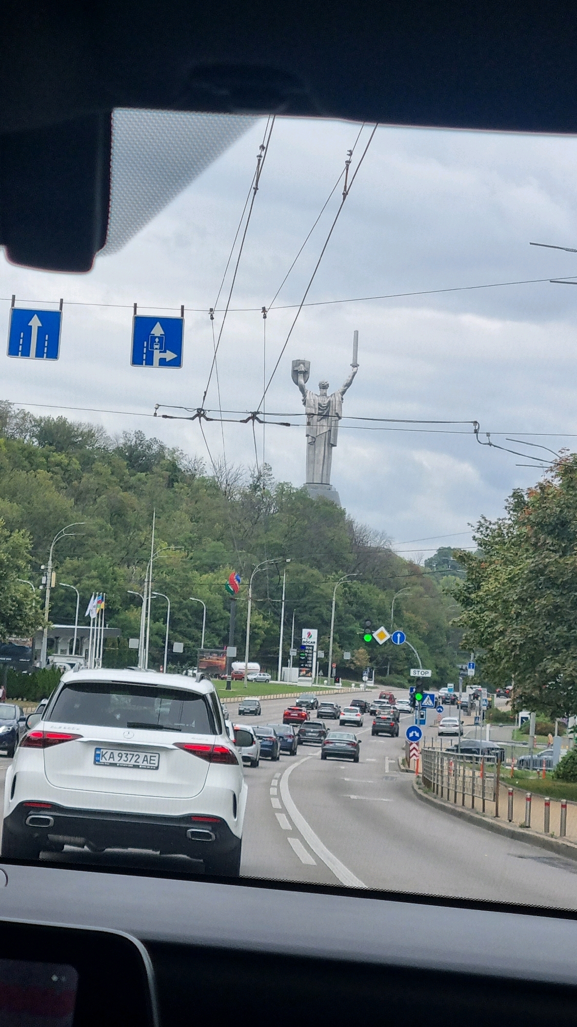 This is a street-level, forward-facing view from within a vehicle traveling on a multi-lane road in Kyiv, Ukraine. The sky is overcast. Prominently featured in the background is the Motherland Monument, a colossal statue of a woman holding a sword and shield, positioned on a hill covered with dense green trees.
In the foreground, a white Mercedes-Benz SUV is visible, with its rear license plate reading "KA 9372 AE". Traffic is moderate, with several cars and SUVs of various makes and models on the road. Traffic lights are visible, with a green light illuminated at one intersection. Road signs are present, including blue directional signs indicating lanes for straight traffic and a right turn, as well as a "STOP" sign.
Along the side of the road, there are streetlights, some with flags attached, including what appears to be the flag of Azerbaijan. There are also gas stations and commercial buildings visible in the distance. Overhead, electrical wires for public transport, likely trams or trolleybuses, crisscross the sky. The scene depicts a typical urban traffic environment in Kyiv.