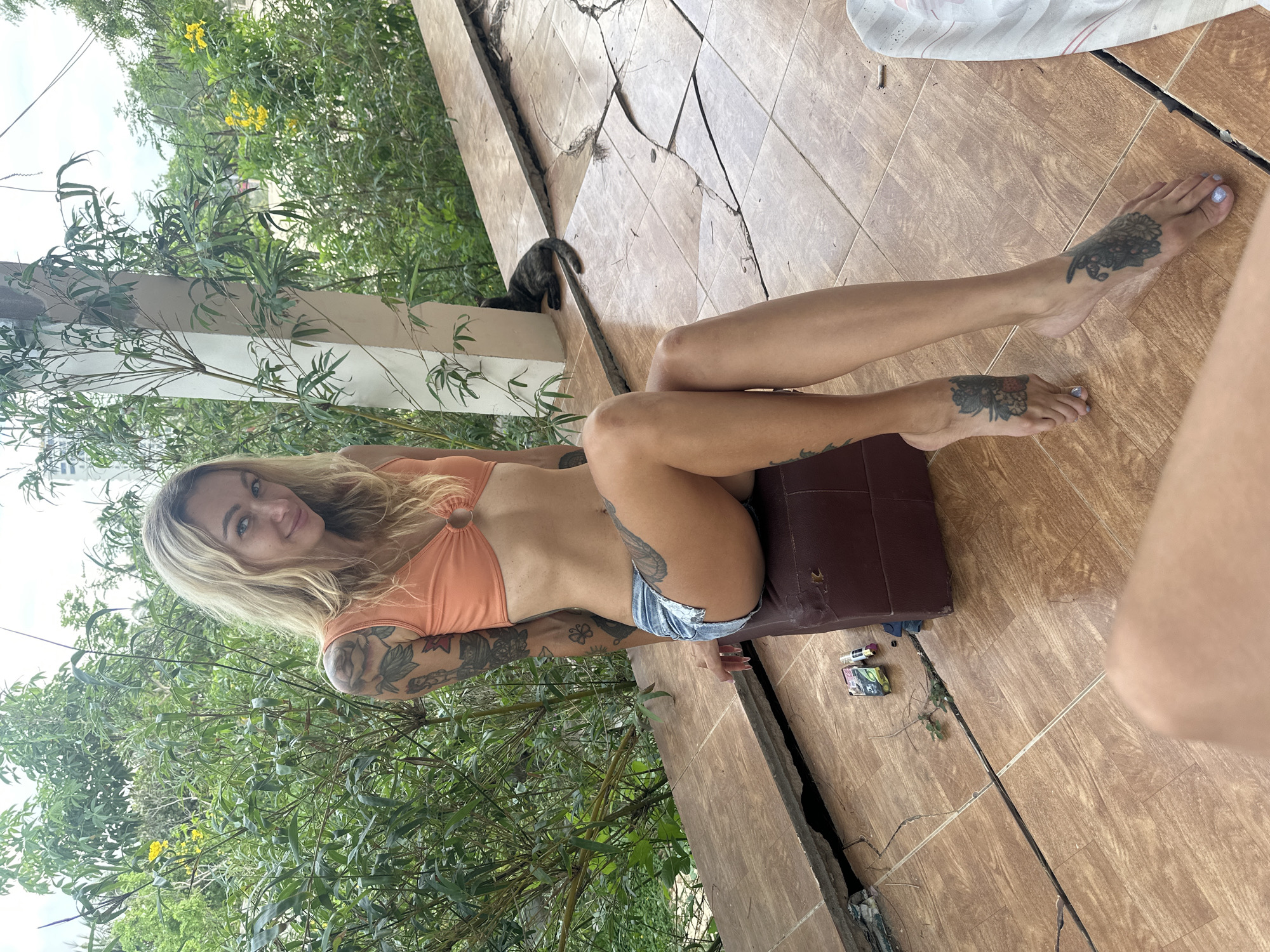 An outdoor, medium shot shows a blonde woman with extensive tattoos lying on a tiled surface, smiling at the camera. She wears an orange bikini top and denim shorts. Her arms and feet are adorned with floral and leaf-patterned tattoos, and her toenails are painted a light purple. She rests on a brown cushioned surface.
The setting is an outdoor patio or balcony in Mũi Né, Vietnam, characterized by a weathered, light brown tiled floor with prominent cracks, particularly along the perimeter where it meets concrete structures. Lush green foliage, including bamboo-like plants and other leafy trees, surrounds the area, with some yellow flowers visible in the background, indicative of a tropical environment. A small, dark-furred cat is visible peeking from behind a concrete pillar in the mid-ground.
Near the woman's cushion, on the cracked tiled floor, are a discarded cigarette butt, a lighter, and a small green and white box, likely a cigarette pack, with some illegible text. The scene is brightly lit, suggesting daytime. The woman appears relaxed and happy.