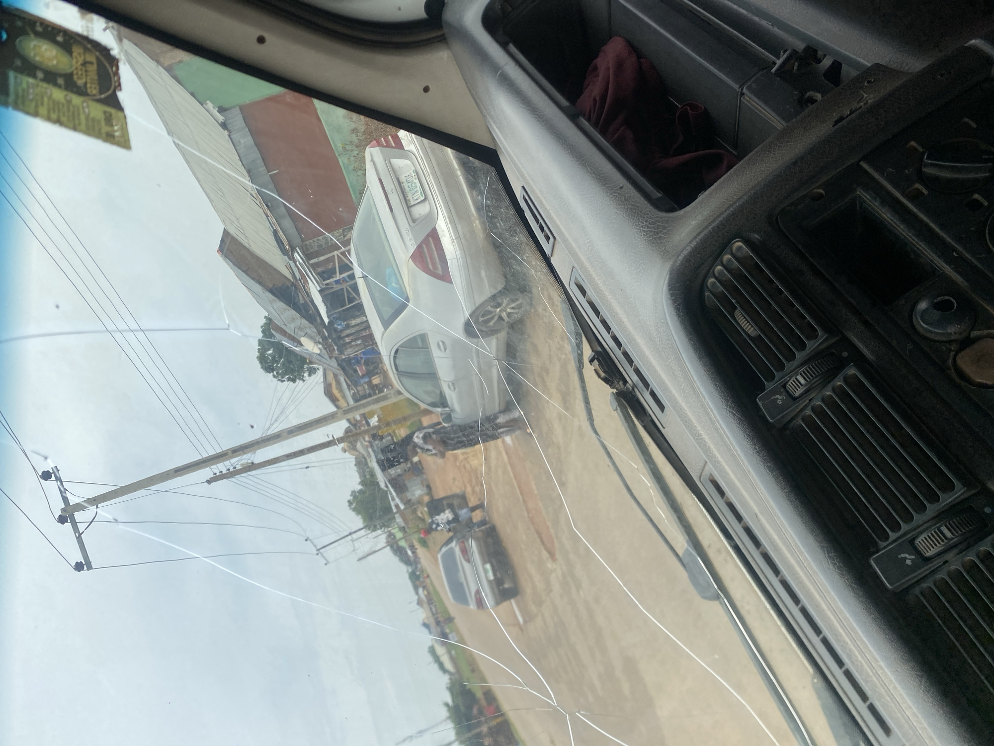 The image is taken from inside a vehicle, looking through a cracked windshield. The scene outside appears to be a dusty road in a Nigerian city, likely DU. There are several cars visible on the road, including a silver Mercedes-Benz sedan with a license plate that reads "KTN 850K". A person in a checkered shirt and dark pants is standing next to one of the cars, and another person in a patterned dress is standing further down the road. Utility poles with numerous wires stretch across the sky, and buildings with corrugated metal roofs line the street. The sky is overcast. The interior of the vehicle shows a dark dashboard with air vents and some controls. A red cloth is visible inside a compartment on the dashboard. The overall impression is of a typical street scene in a Nigerian town or city.