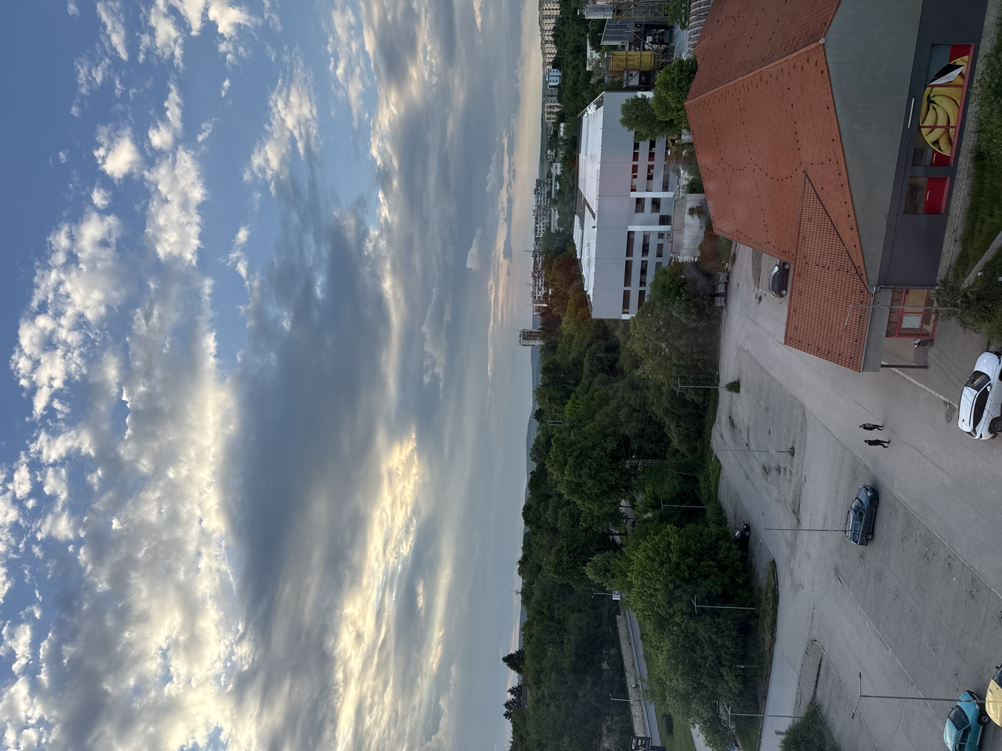 The image depicts an aerial view in Lovech, Bulgaria. The sky is predominantly filled with clouds, exhibiting a mix of white and grey tones, with hints of blue sky visible. The landscape below the sky includes a blend of urban structures and natural elements. Buildings with orange tiled roofs and white walls are visible, along with dense green trees. A parking lot occupies a significant portion of the scene, with several cars parked. Two people are visible on the pavement.