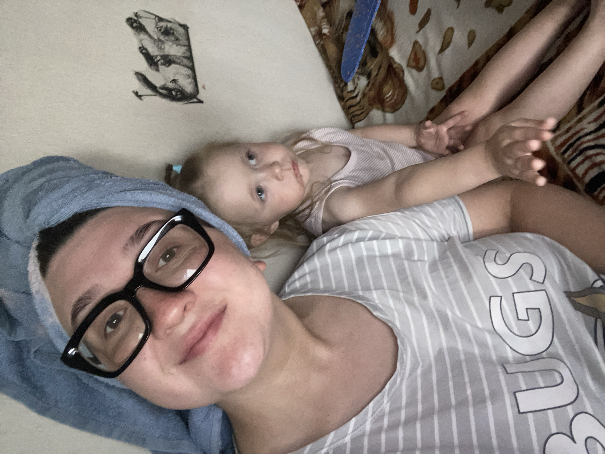 In an indoor setting, a woman wearing a blue towel on her head and black-rimmed glasses poses for a selfie, her face partially obscured. She is wearing a grey and white striped t-shirt with the word "BUGS" visible. Beside her, a young child with blonde hair is looking away, wearing a light pink striped tank top. The background includes a light-colored wall with a drawing of an animal and a patterned bedspread. The overall scene suggests a casual, domestic moment. This image was taken in the city of Chaikovsky, Russia.