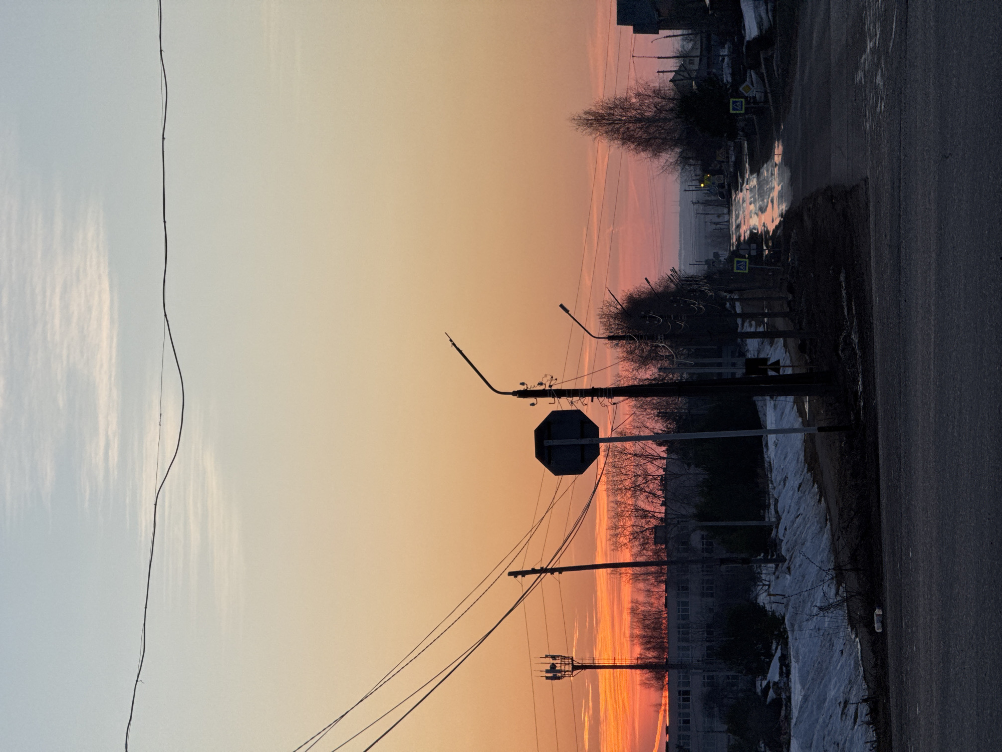 The photo shows a street scene in Прикамский, Russia, during a sunset or sunrise, with the sky exhibiting colors of orange, pink, and pale blue. The street, paved with asphalt, appears to be wet, possibly from melting snow. Along the roadside, remnants of snow are visible, indicating a cold climate. Streetlights and utility poles line the road, and an octagonal stop sign is prominently placed. In the background, multi-story buildings and bare trees are silhouetted against the colorful sky. A network of electrical wires spans across the scene. The image is oriented sideways, with the sky to the left and the road to the right.