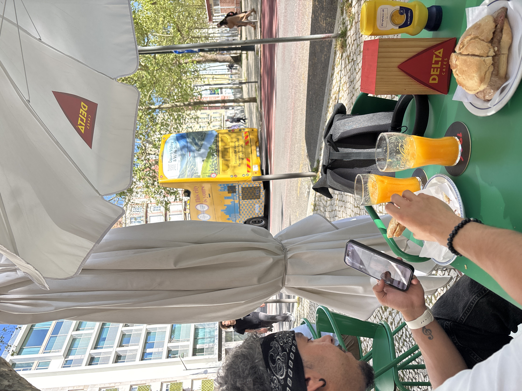 The image depicts a man seated at an outdoor cafe, holding a smartphone while taking a bite of food. On the table are two glasses of orange juice, a half-eaten sandwich on a plate, a backpack, a mustard bottle, and a wooden box with the "DELTA CAFÉS" logo. An advertising sign for "Douro Valley & Wine Tour" is visible in the background, along with a view of a street in Lisbon, Portugal, with people walking by. The cafe is shaded by a white umbrella that also carries the "DELTA" logo. The man is wearing a black bandana, a white t-shirt, and a watch on his left wrist. His jeans are visible. There is a tattoo of a music clef on his left wrist. The restaurant table is green and the chairs are also green metal.