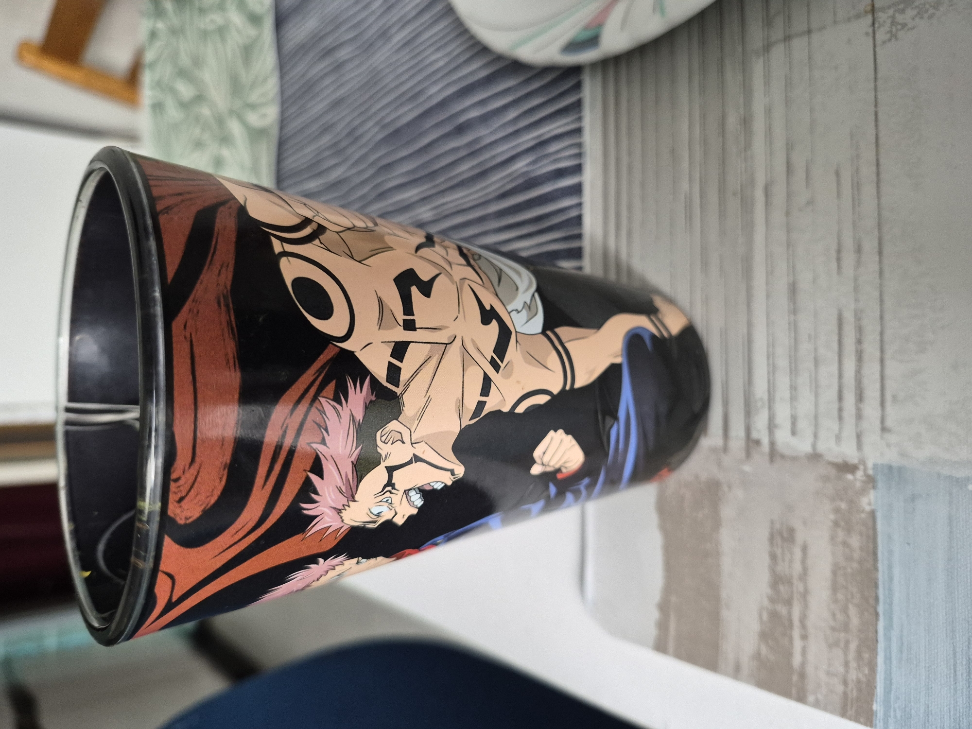 A close-up, eye-level shot shows a black glass with a depiction of a character from the anime Jujutsu Kaisen. The character has pink spiky hair, blue eyes, and is shown with a muscular physique and tattoos. The glass is placed on a table with a blue and white striped placemat, and a light gray and blue striped runner. In the background, a portion of a light blue chair is visible, and a blurred wooden chair is present. The lighting is natural.