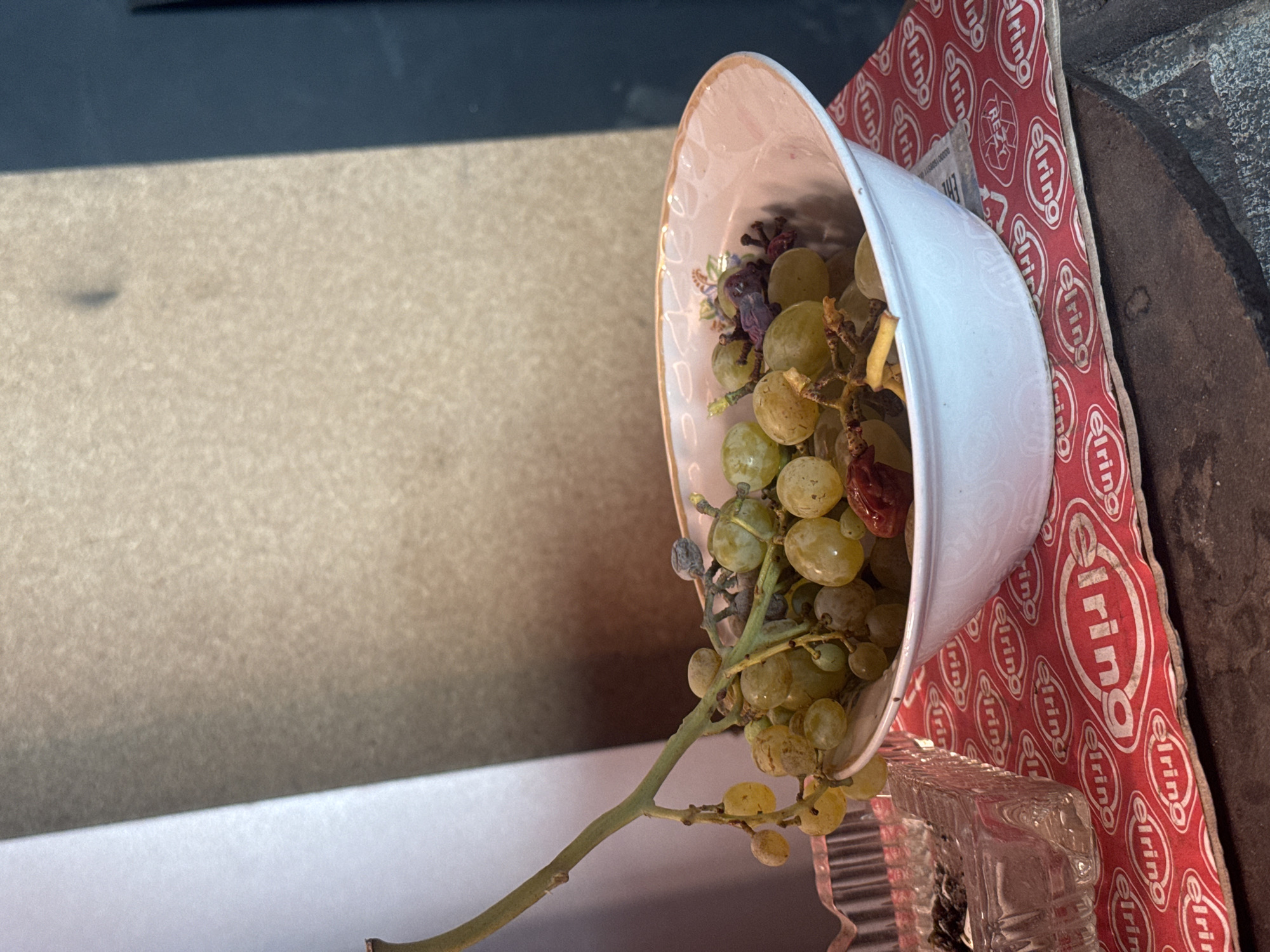 A close-up, eye-level shot captures a white ceramic bowl filled with light green grapes and some dried fruits. The bowl, decorated with a floral pattern around its rim, sits on a red placemat with a repeating "elring" logo. To the left, a clear glass ashtray contains cigarette butts. The background consists of a textured, light brown wall and a dark vertical surface, casting shadows. The lighting is dim, suggesting an indoor setting. The scene appears to be a still life, likely arranged in a home or a small shop, possibly in Baku, Azerbaijan, given the presence of the "elring" logo which is a brand of automotive parts and services, commonly found in markets and workshops. There are no people or animals present.