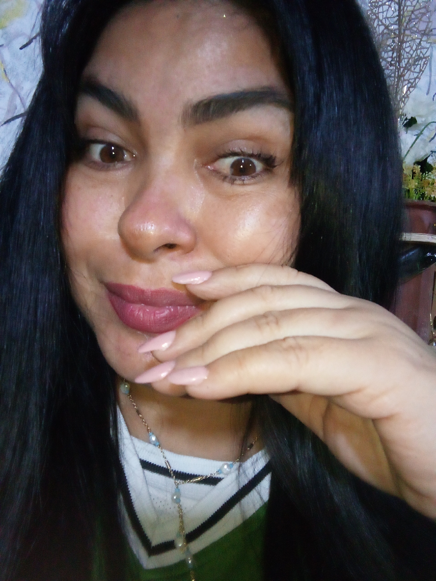 A woman with dark hair is facing the camera, her right hand partially covering her mouth. She has brown eyes and light pink lipstick. A necklace with light blue beads is visible at her neck. She is wearing a white and green striped shirt. There is a tear on her left cheek. The background is blurred, suggesting an indoor setting. There is no discernible information in the image to indicate the location is specifically Juan Griego, Venezuela.