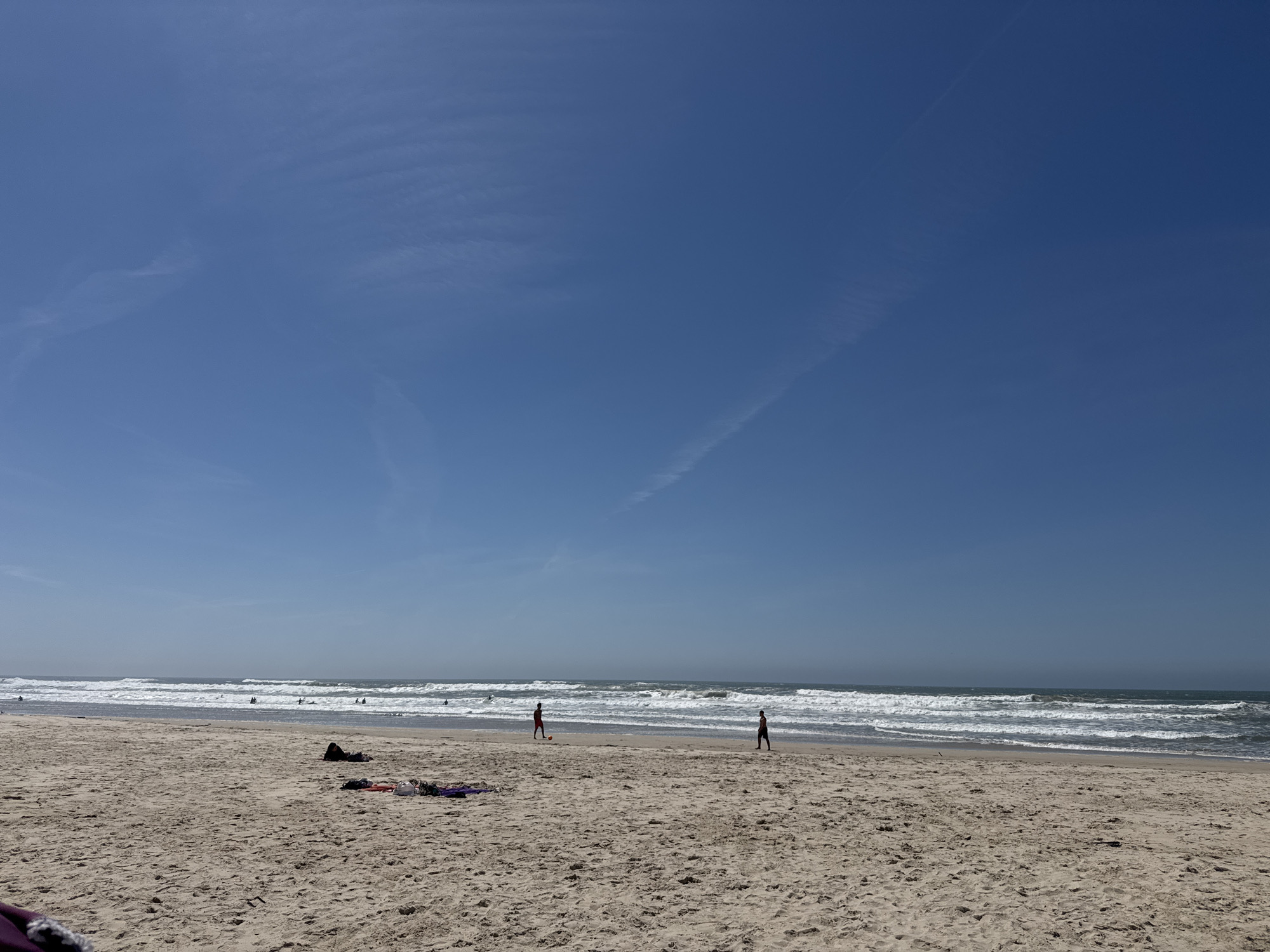 The image depicts a beach scene in Figueira da Foz, Portugal on a sunny day with a clear blue sky. The sandy beach occupies the lower half of the image, showing footprints and small objects scattered across the sand. Two figures are visible on the beach: one person walks towards the sea, and another is closer to the water, possibly kicking a red ball. The ocean stretches across the middle of the image, with white-capped waves rolling towards the shore. In the background, several other people can be seen in the water, possibly surfing or swimming. The sky is a clear blue with faint contrails.