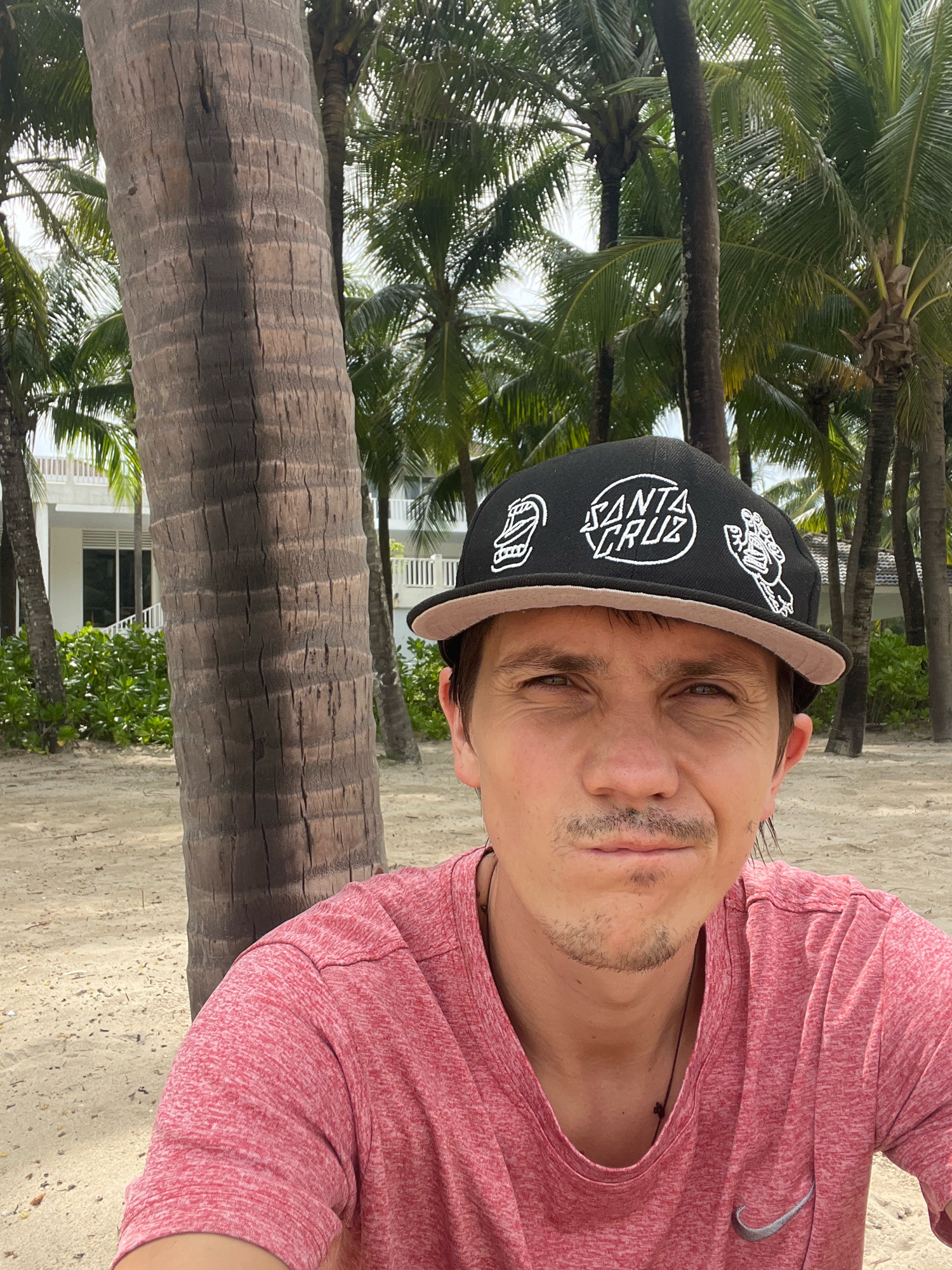 A man is taking a selfie on a beach in Đà Nẵng, Vietnam. He is wearing a red t-shirt and a black baseball cap with white graphics. He has a mustache and a goatee. Behind him, there are palm trees and a white building. The beach appears to be sandy, and the weather looks sunny. The man is leaning against a palm tree.