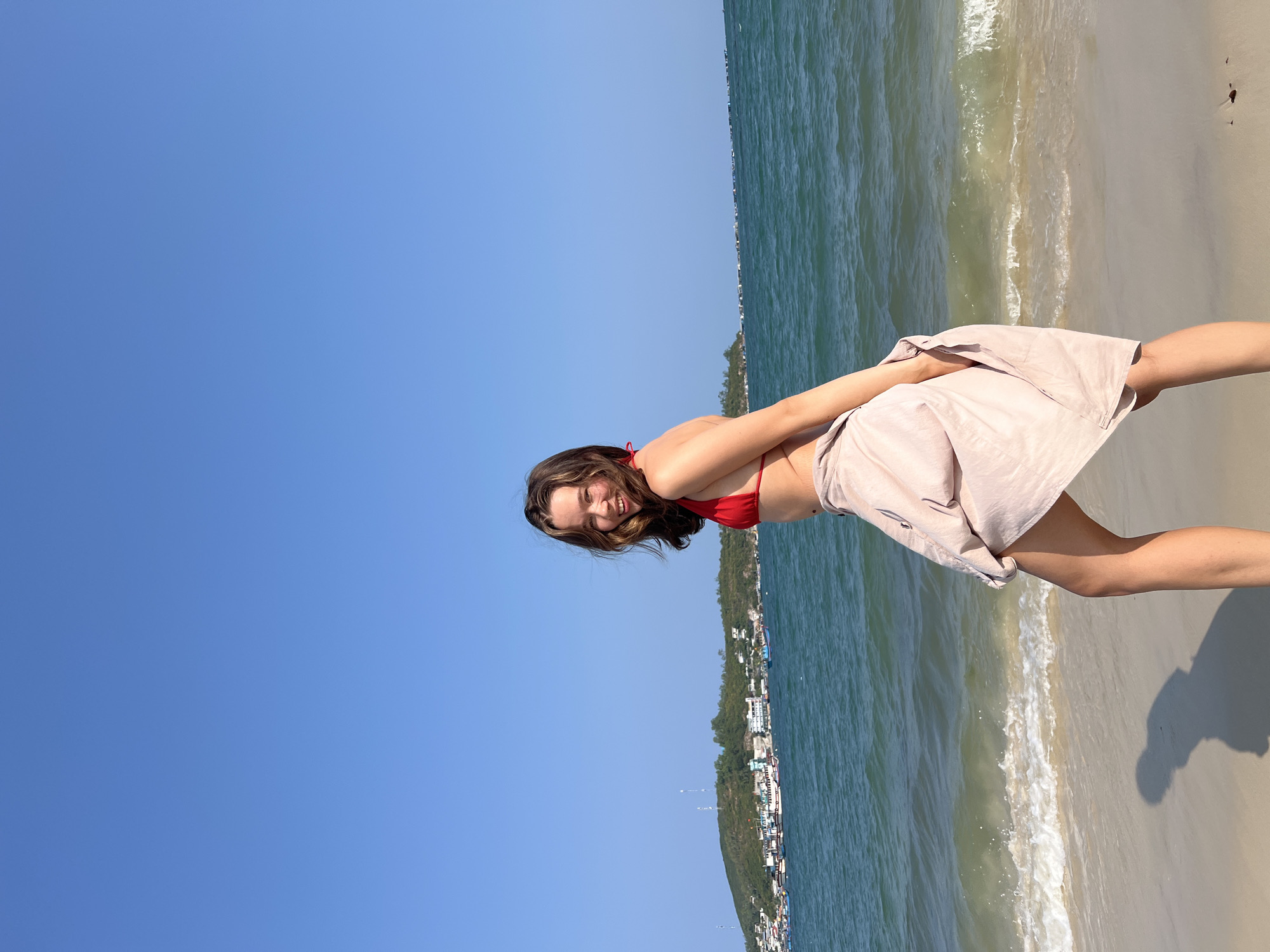 A young woman in a red bikini top and light pink skirt stands on a sandy beach on a bright, sunny day. Her body is rotated slightly, and she is smiling. Behind her, the turquoise water of the ocean meets the sand. In the distance, a hillside dotted with buildings is visible, likely part of the urban area of Nam Nha Trang, Vietnam. The sky is clear blue.